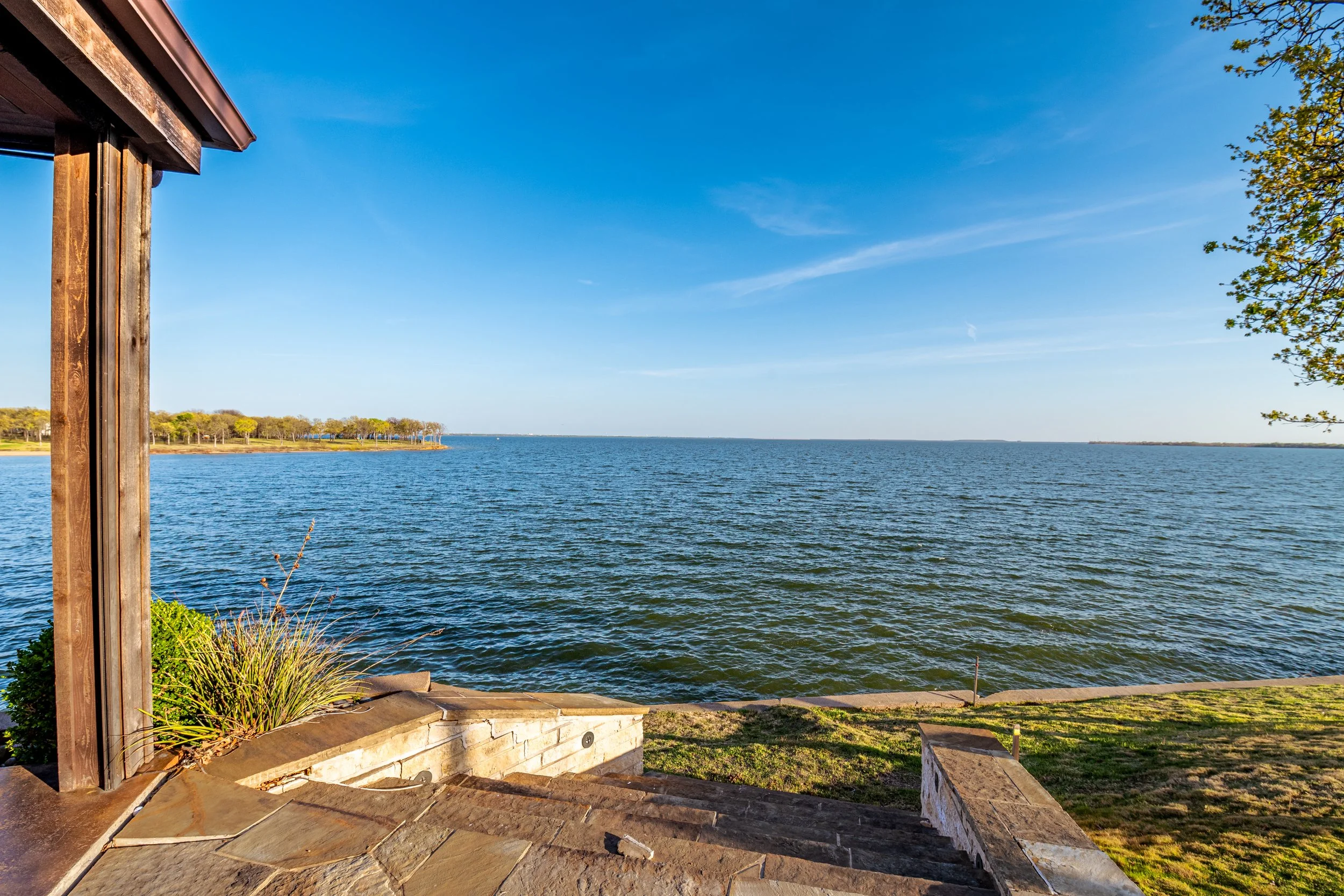 View of a large body of water with trees in the distance, seen from a stone patio with a wooden post and some greenery in the foreground, under a bright blue sky with a few wispy clouds