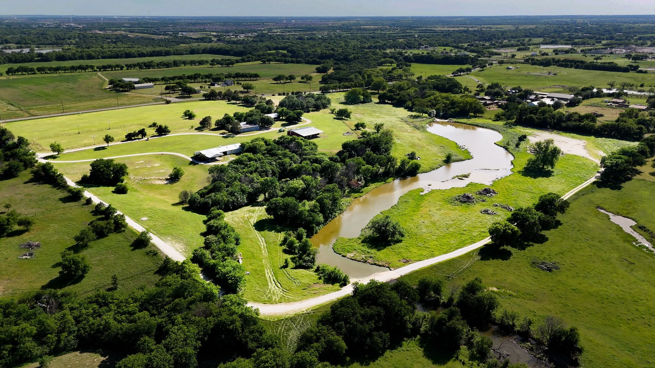 Aerial view of a rural landscape with green fields, trees, winding river, and several farm buildings. Wide open spaces and a distant city skyline on the horizon.
