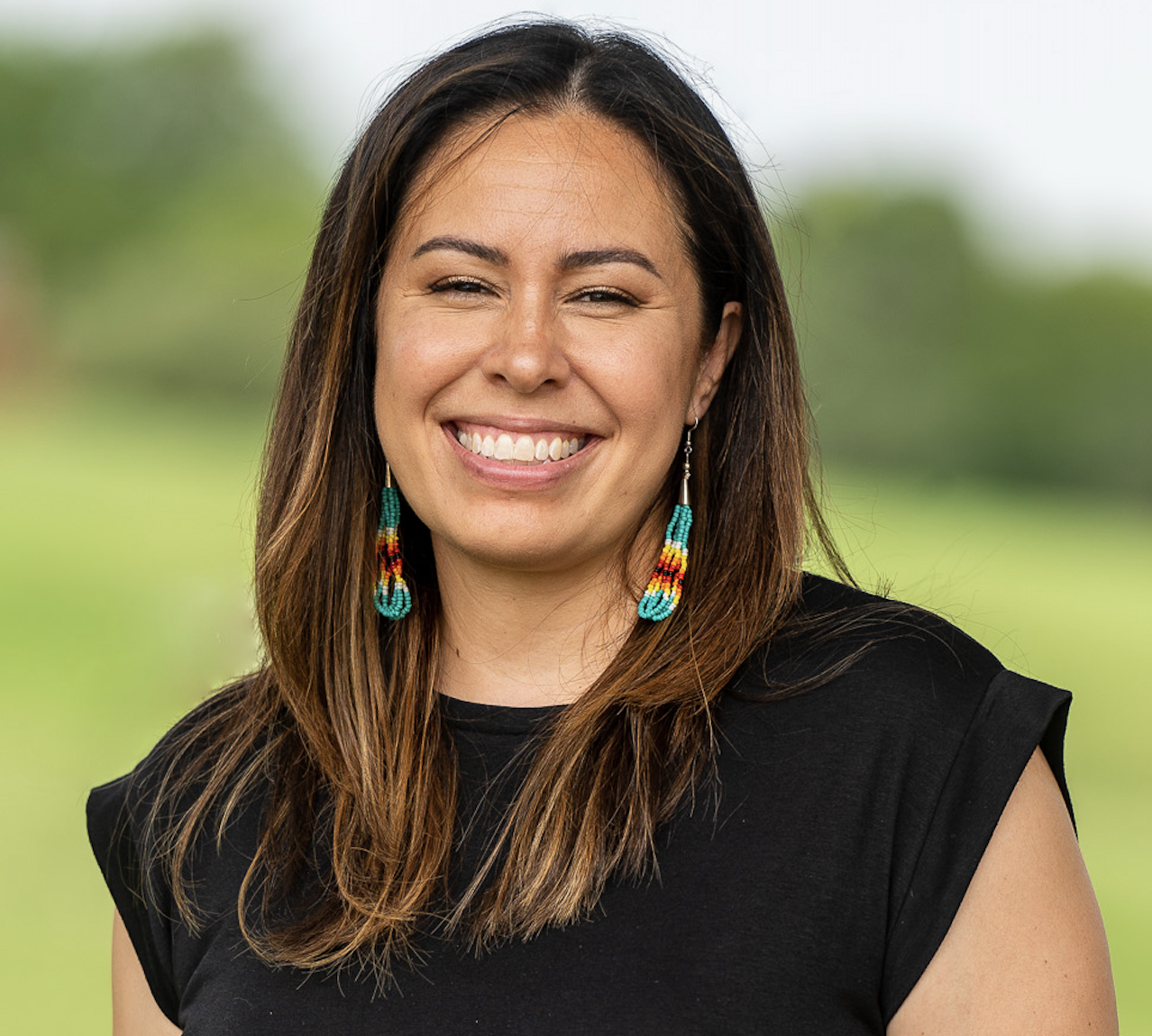 A woman smiling outdoors with a blurred green background, wearing a black sleeveless top and colorful beaded earrings.