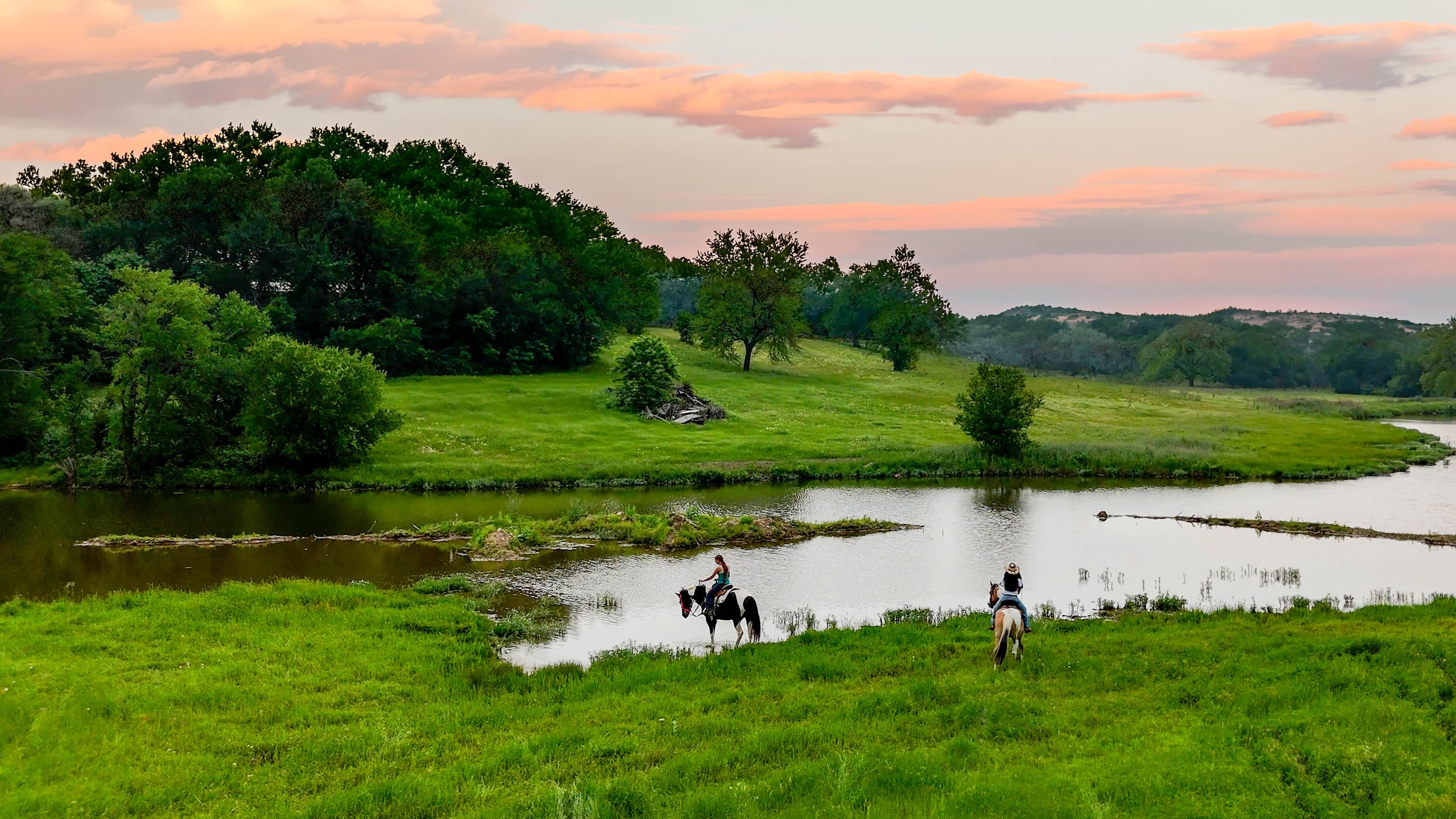 People horseback riding along the edge of a river in a lush green landscape during sunset, with trees and rolling hills in the background.
