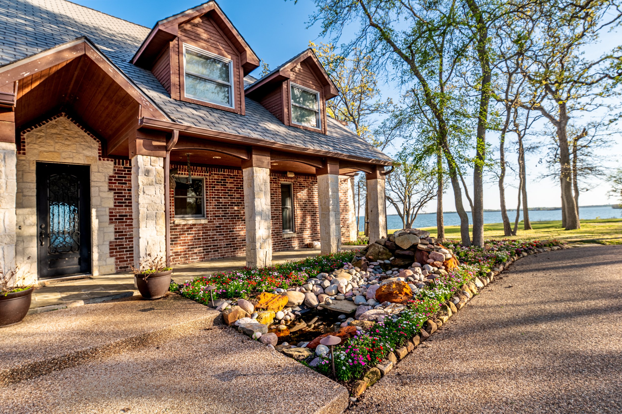 A brick house with a stone and brick exterior, large windows, and a covered porch supported by stone columns. The house is surrounded by colorful flowers, rocks, and a gravel pathway. In the background, there are tall trees and a body of water.