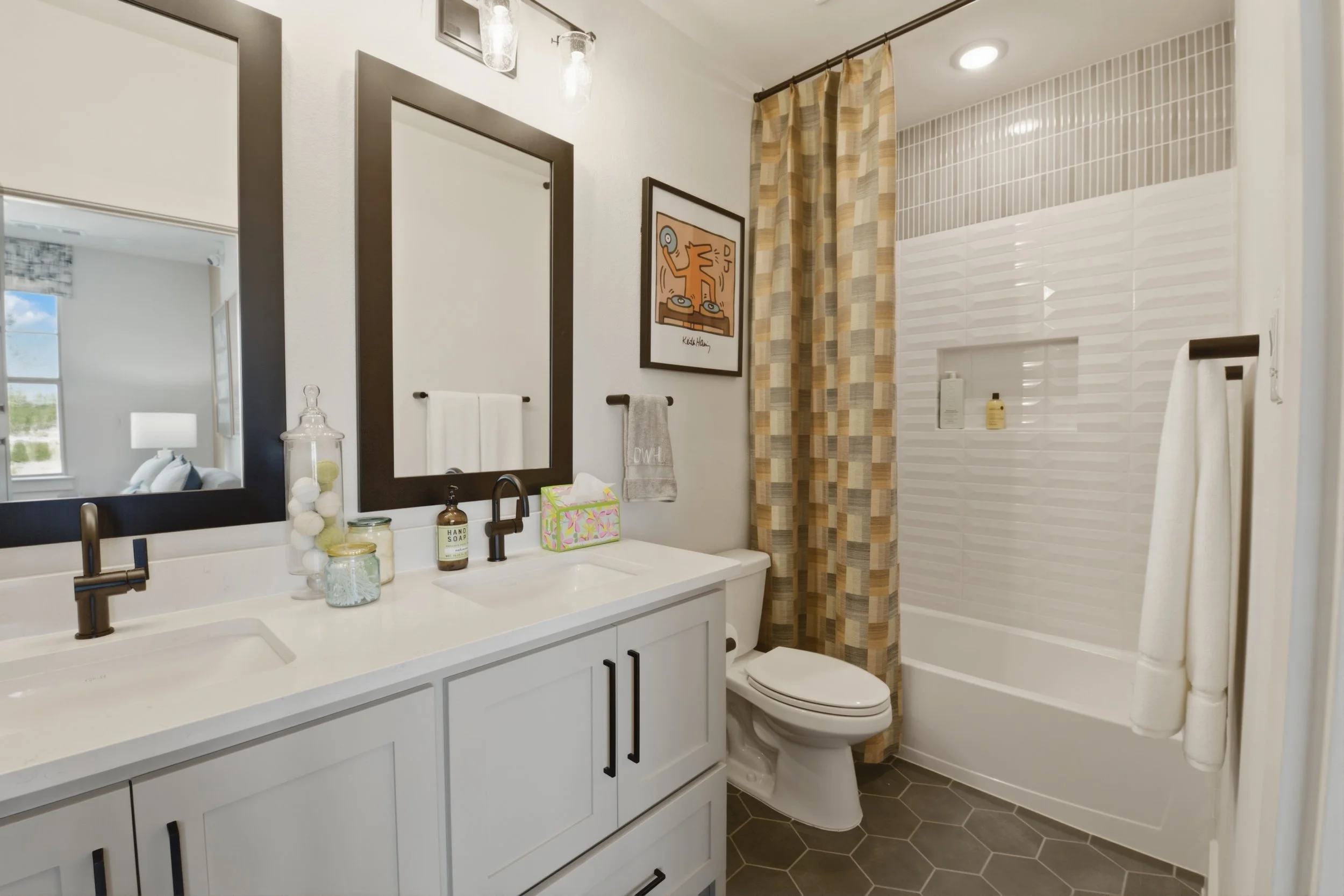 A modern bathroom featuring a white vanity with a double sink, large mirrors, and black cabinet handles. Decor includes a glass container filled with decorative orbs, a tissue box, and two soap dispensers. To the right, there's a shower area with pat