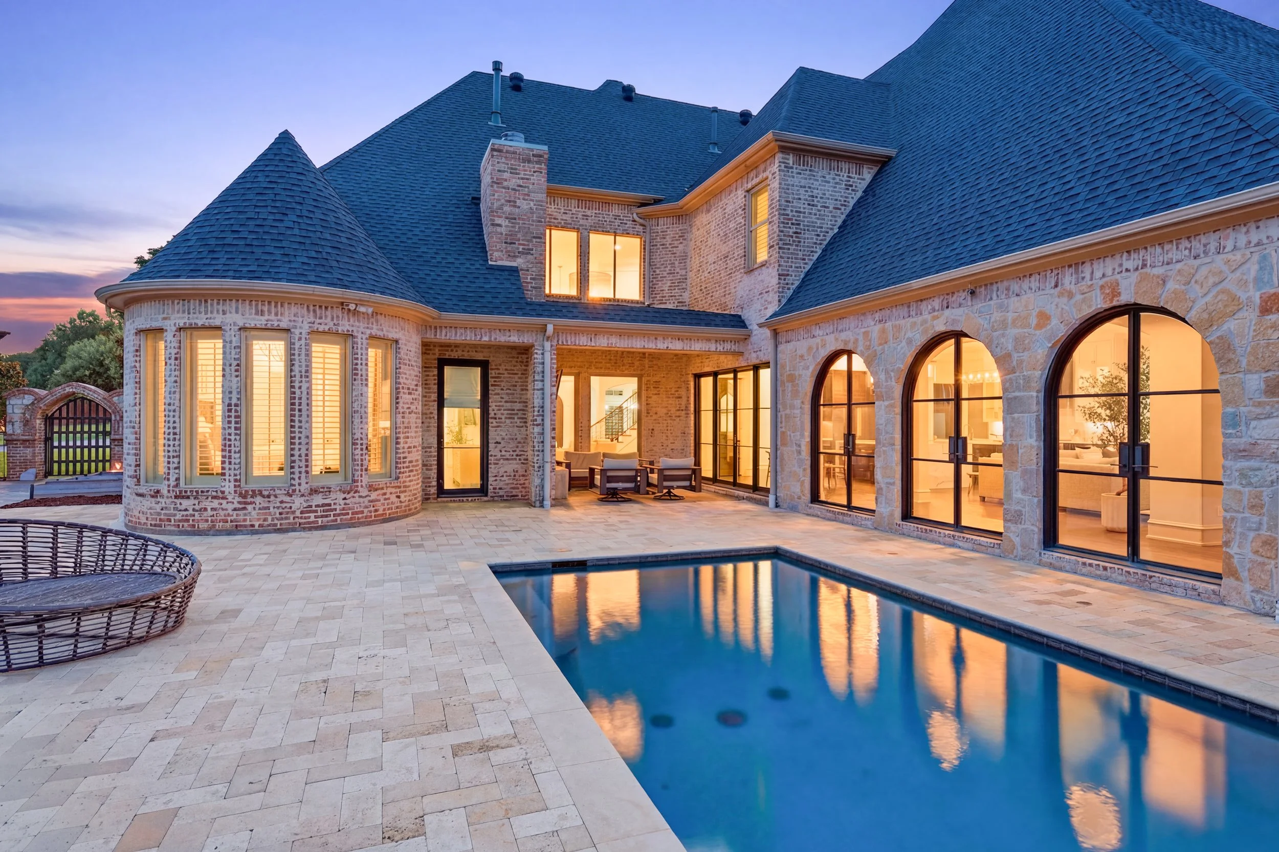 The image shows the backyard of a large brick house at twilight with illuminated windows. There is a pool in the foreground reflecting the sky, surrounded by a stone-tile patio. Outdoor furniture is visible near the house.