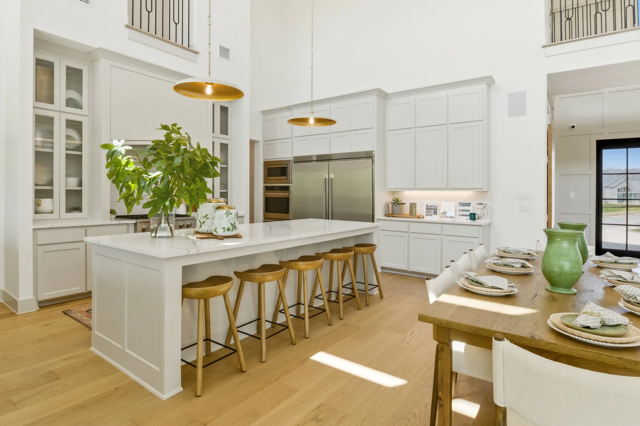 A spacious kitchen and dining area with white cabinetry, a large island with barstools, hanging gold pendant lights, and a dining table set with plates, napkins, and green vases. There is a large plant on the island and built-in stainless steel appli