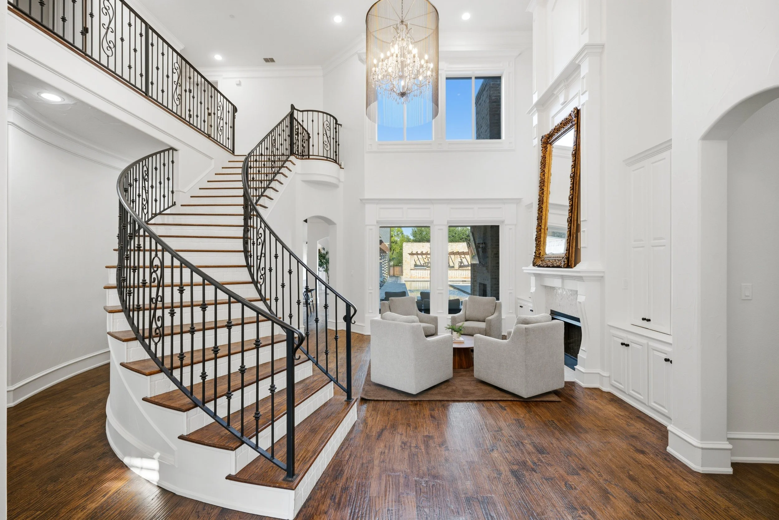 Elegant living room with white walls, a staircase with black wrought iron railing, a chandelier, large windows, and a sitting area with several chairs.