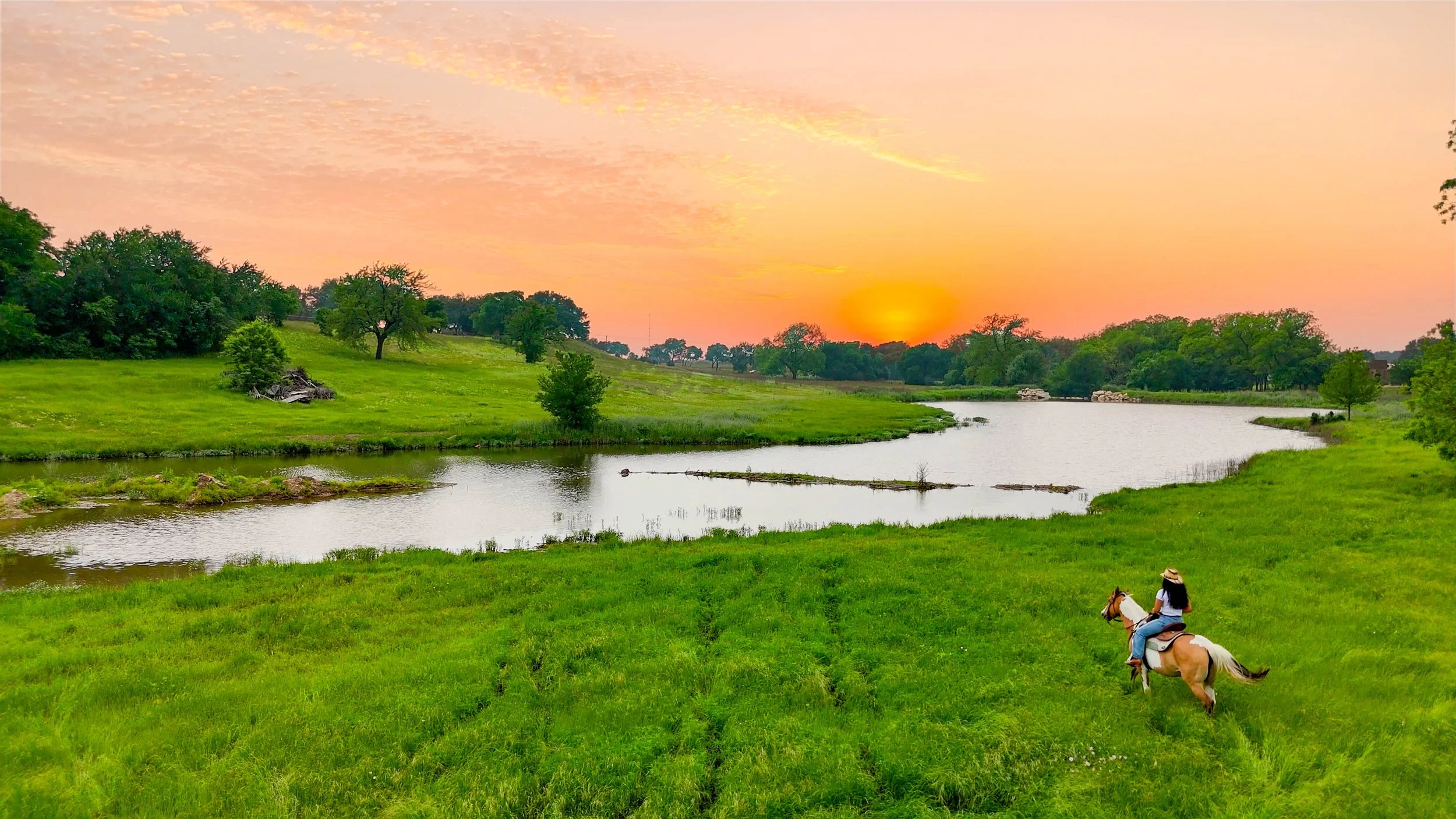 A person riding a horse on a grassy field near a river during sunset, with trees in the background and a partly cloudy sky.