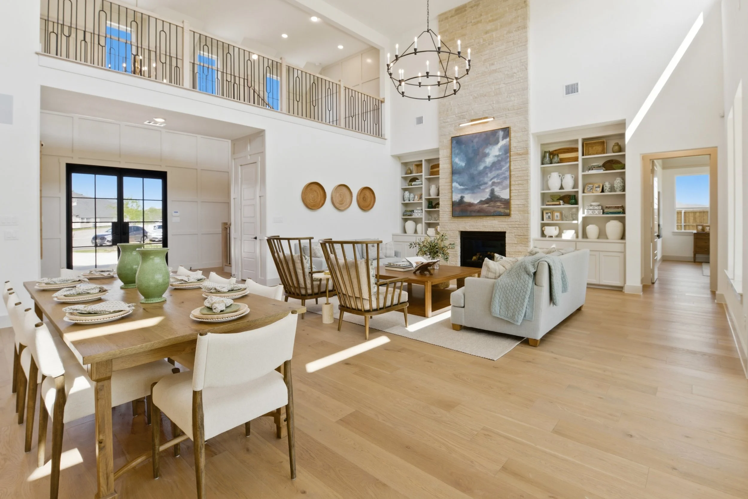 Bright and spacious living room with a high ceiling, large chandelier, a fireplace with a painting above, built-in shelves with decorative items, and a dining table set with plates and green vases.