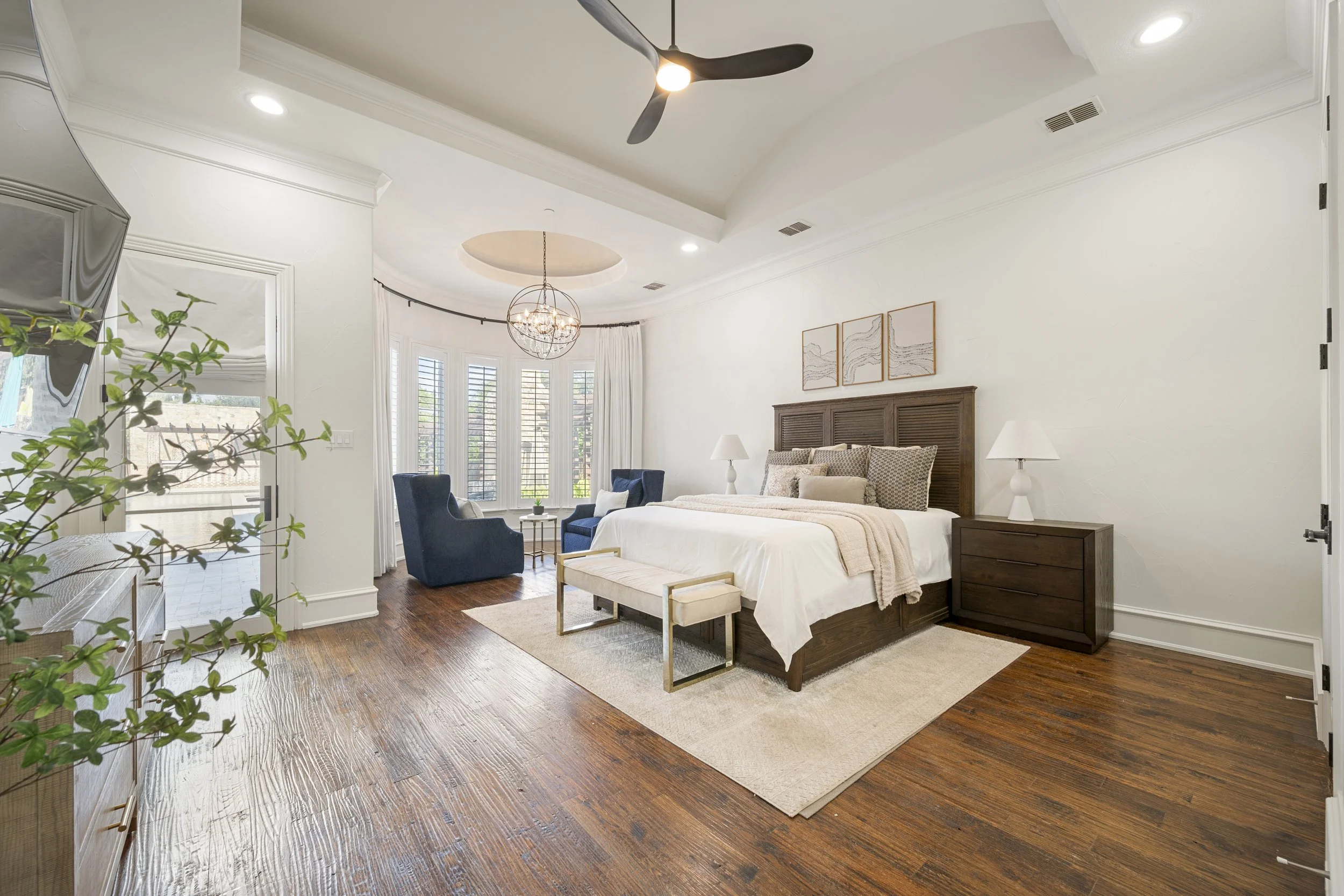 A spacious bedroom with a large bed, dark wood headboard, two white bedside lamps, and a bench at the foot of the bed. The room features a curved window with white blinds, a chandelier, dark blue armchairs, and hardwood floors with a small rug.
