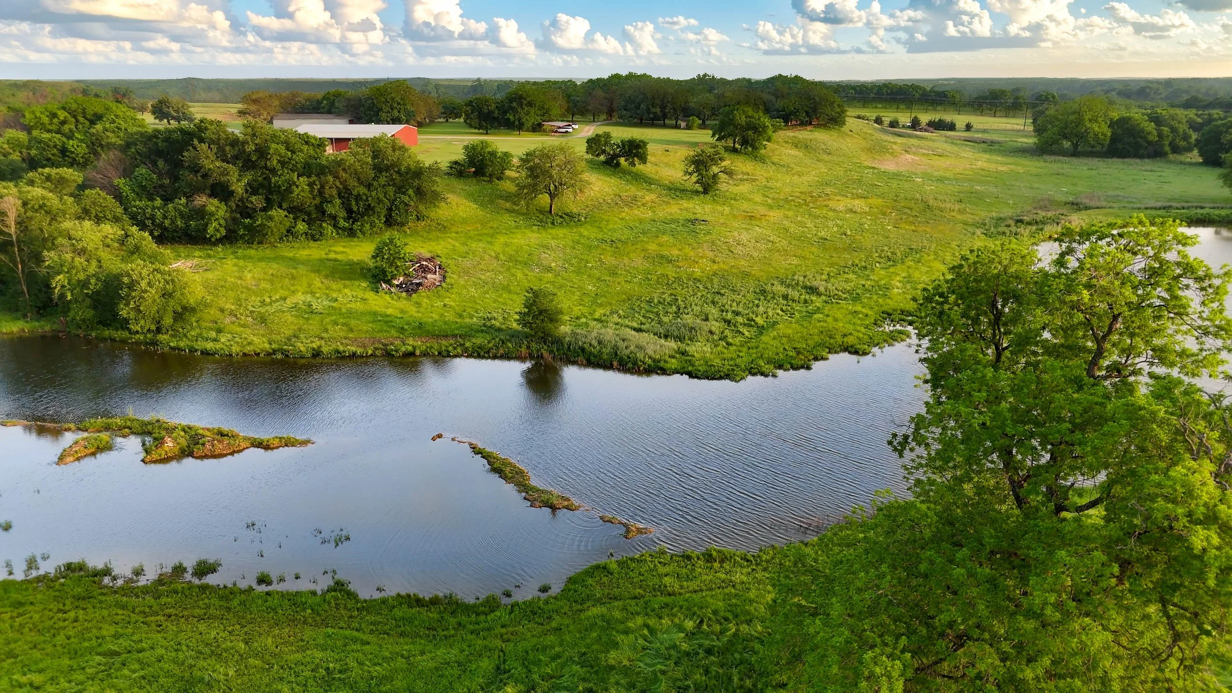 A scenic view of a river flowing through a green landscape with trees, grassy fields, and farm buildings in the distance under a partly cloudy sky.