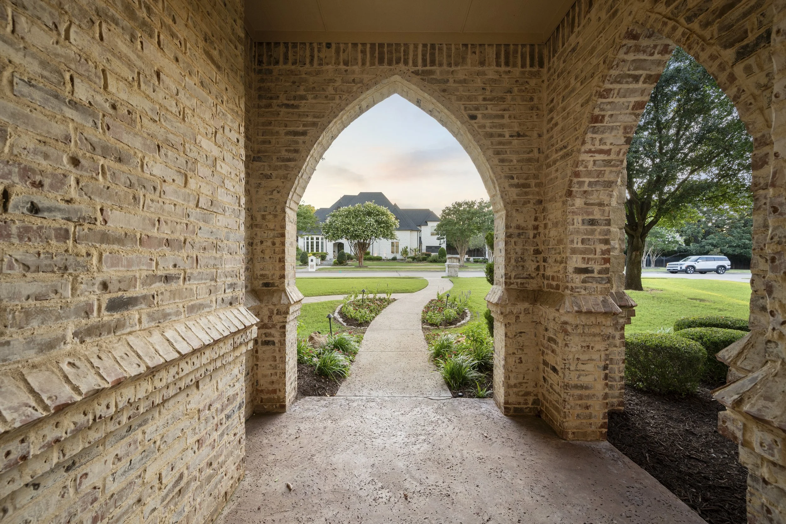 View of a suburban neighborhood through arched brick openings, showing a sidewalk, greenery, houses, and parked cars.