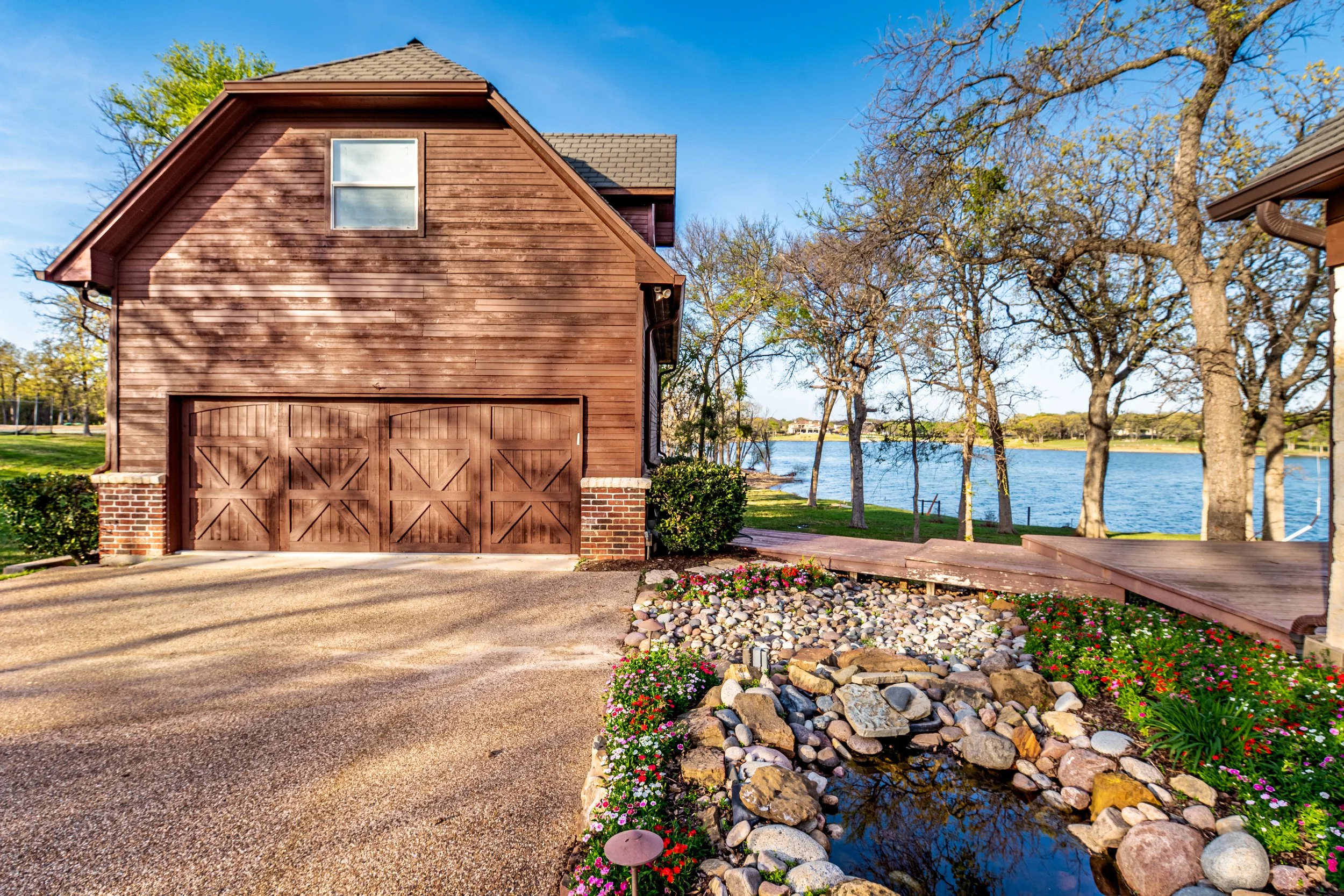 A wooden garage and house with a sloped roof, next to a body of water, surrounded by trees and a flower garden.