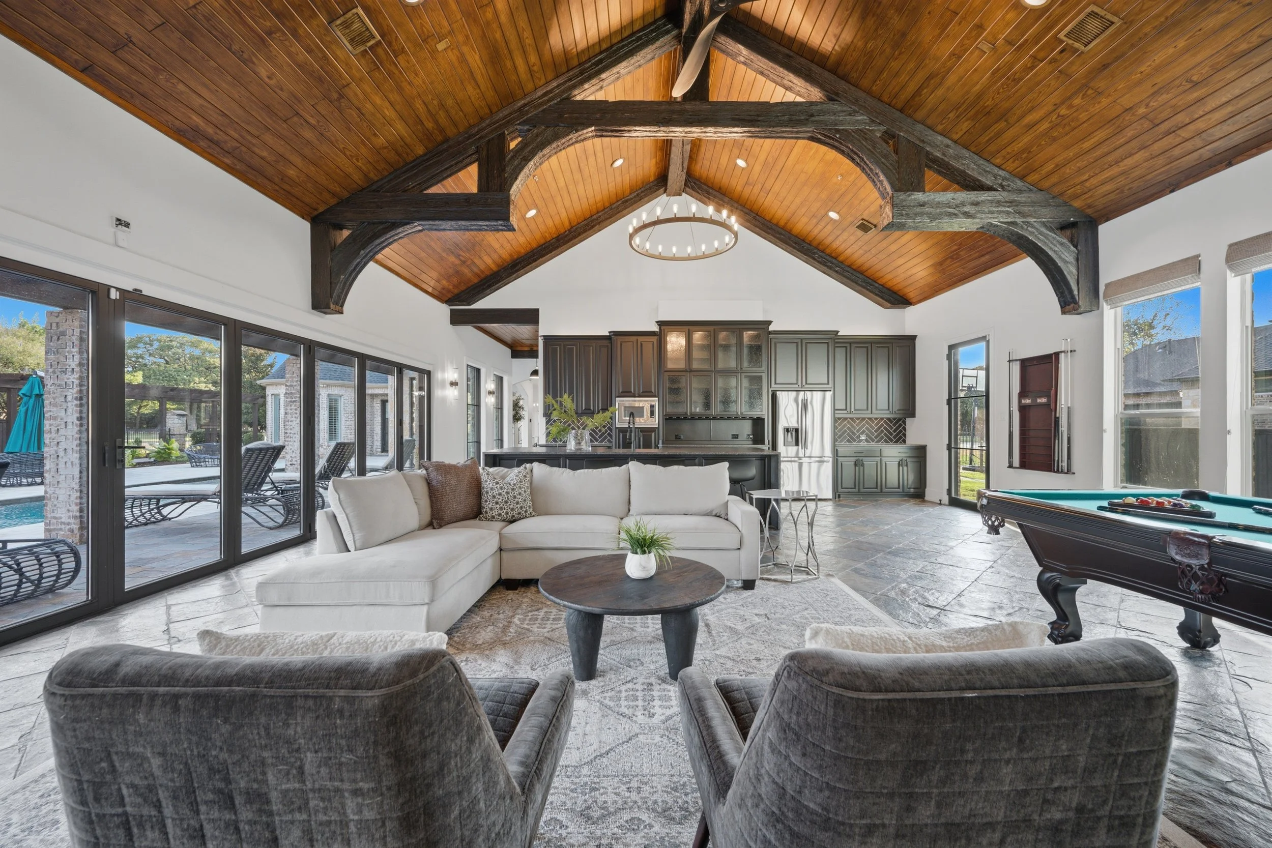 Living room with vaulted wood ceiling, dark wood beams, large windows, light-colored sofa, pool table, and outdoor patio visible through glass doors.