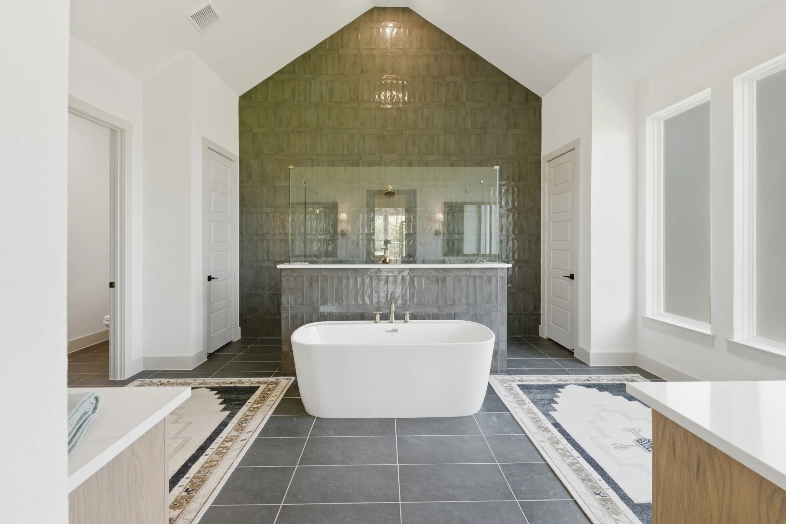 Modern bathroom with a white freestanding bathtub in front of a black tile wall with a glass shower enclosure. White walls with windows and black door handles.