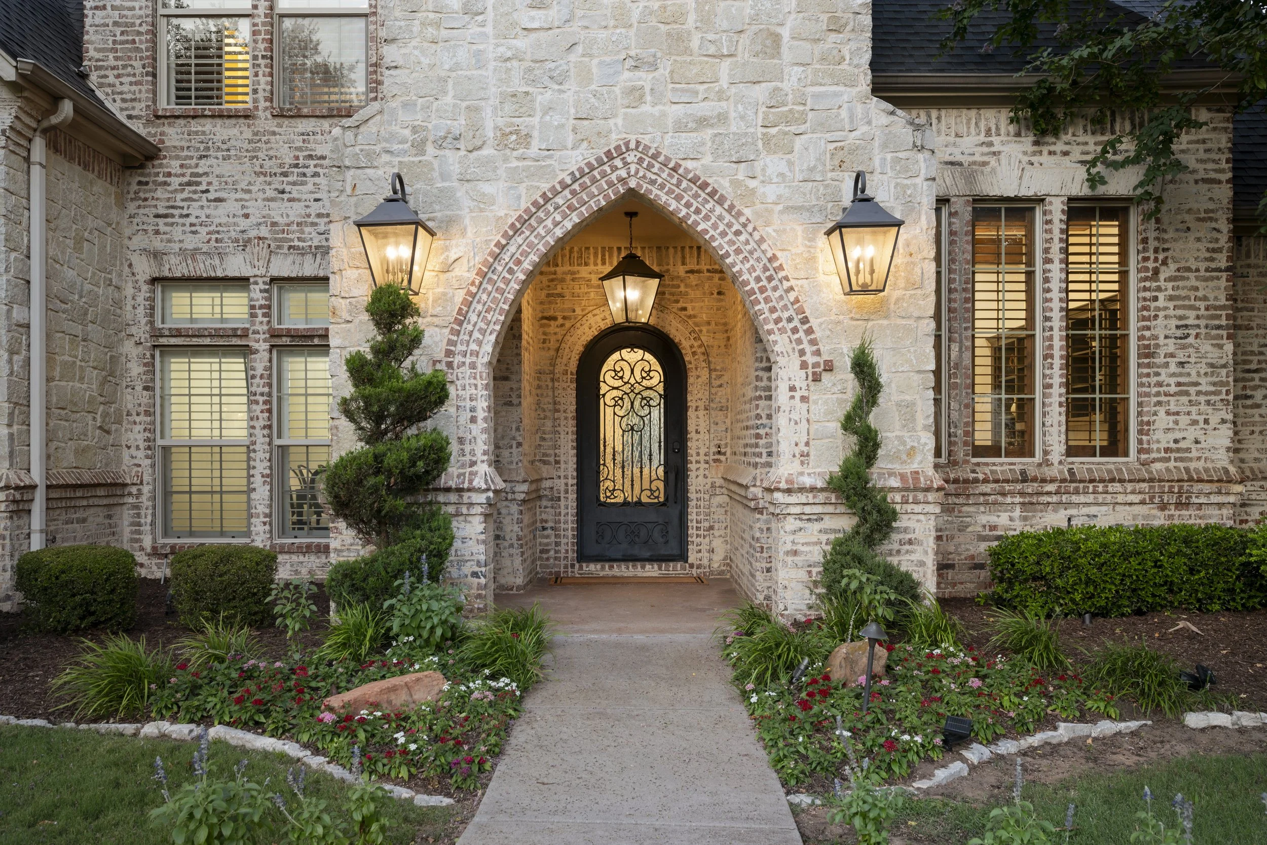 Front view of a brick house with an arched entrance, decorated with black wrought iron gate and three hanging lanterns, surrounded by a small garden with bushes, flowers, and a concrete pathway.
