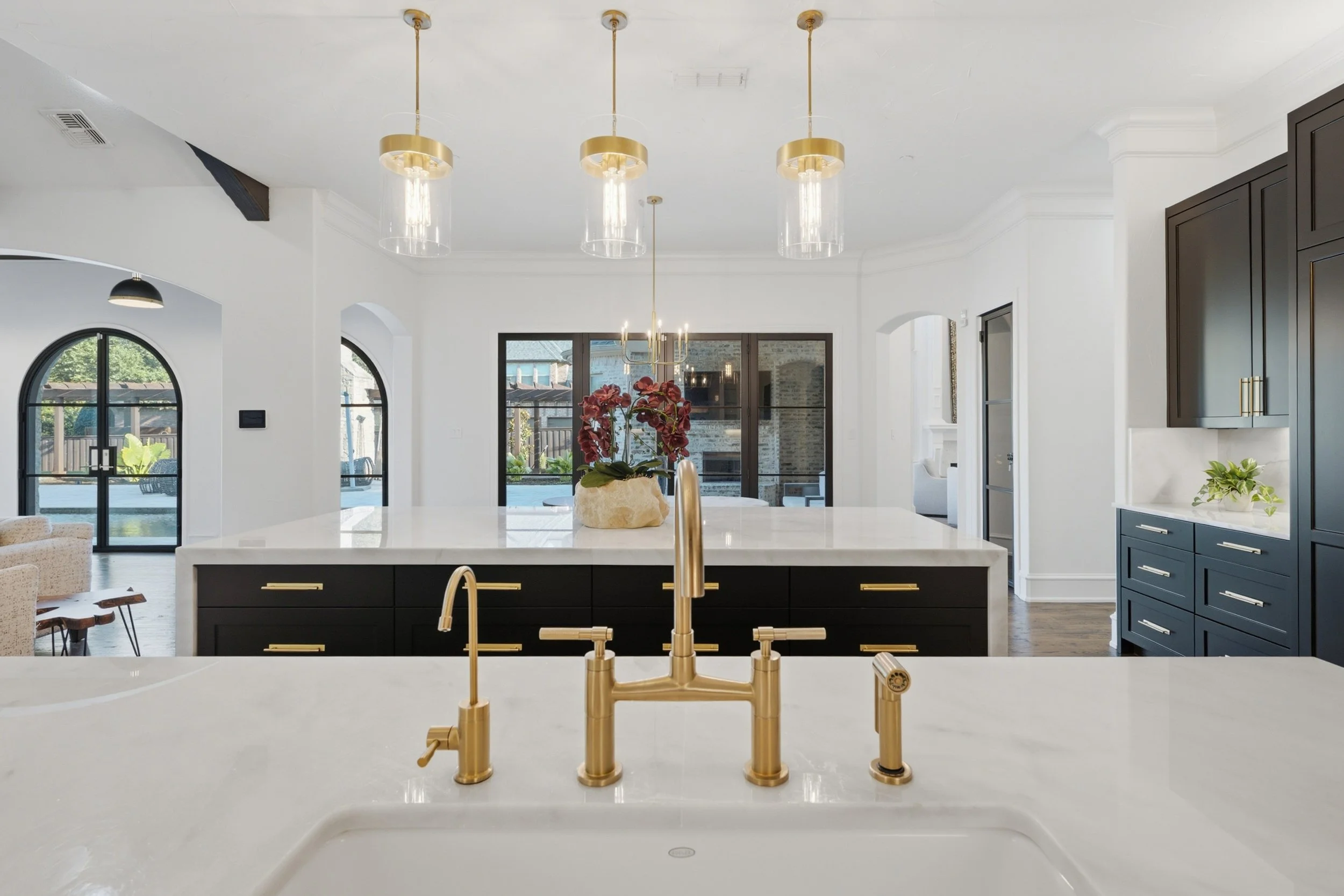 Modern kitchen with a white marble island, gold faucet, and black cabinetry, bright open space with large windows, pendant lights hanging from the ceiling.