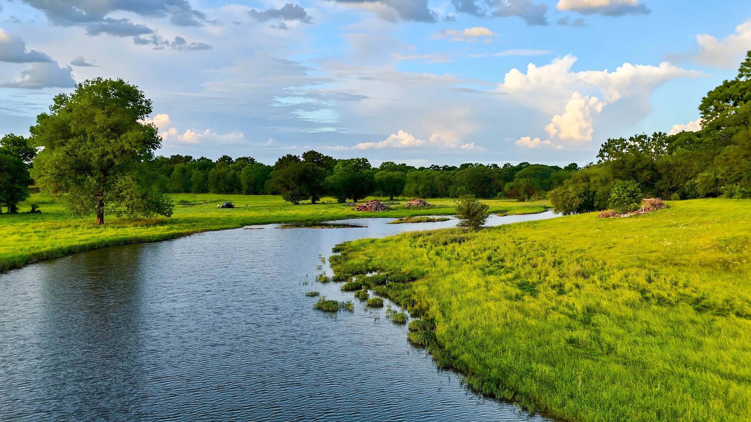 A peaceful landscape featuring a river flowing through lush green fields and trees under a partly cloudy sky.
