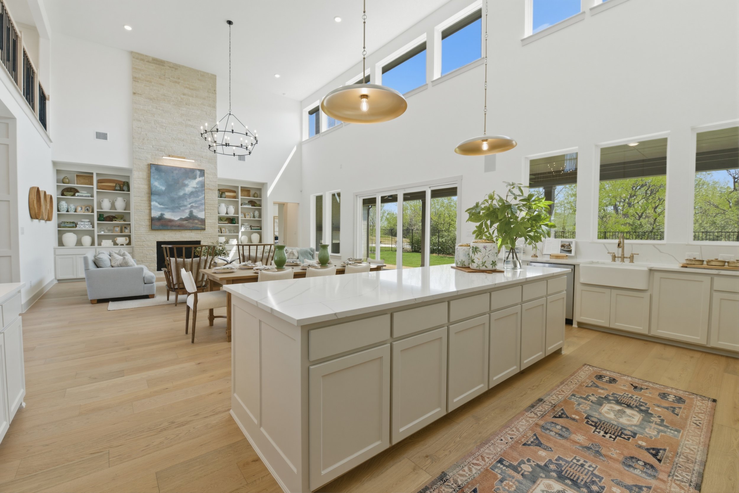 Open-concept kitchen and living room with high ceilings, large windows, wood flooring, white cabinetry, and a stone fireplace with built-in shelves. Decor includes plants and artwork, with a view of greenery outside.