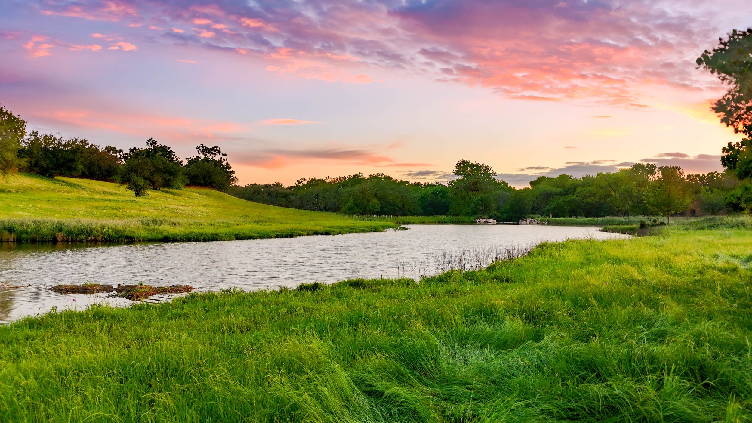 Scenic view of a river flowing through lush green grass and trees with a colorful sunset sky overhead.