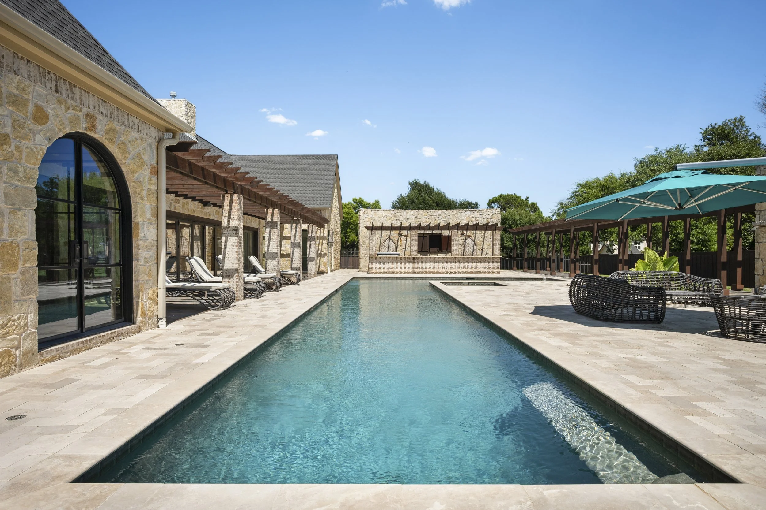 A backyard with a swimming pool, surrounded by stone patio, lounge chairs, and a covered sitting area with a green umbrella. A stone house with large windows and a wooden pergola is visible in the background under a blue sky with a few clouds.
