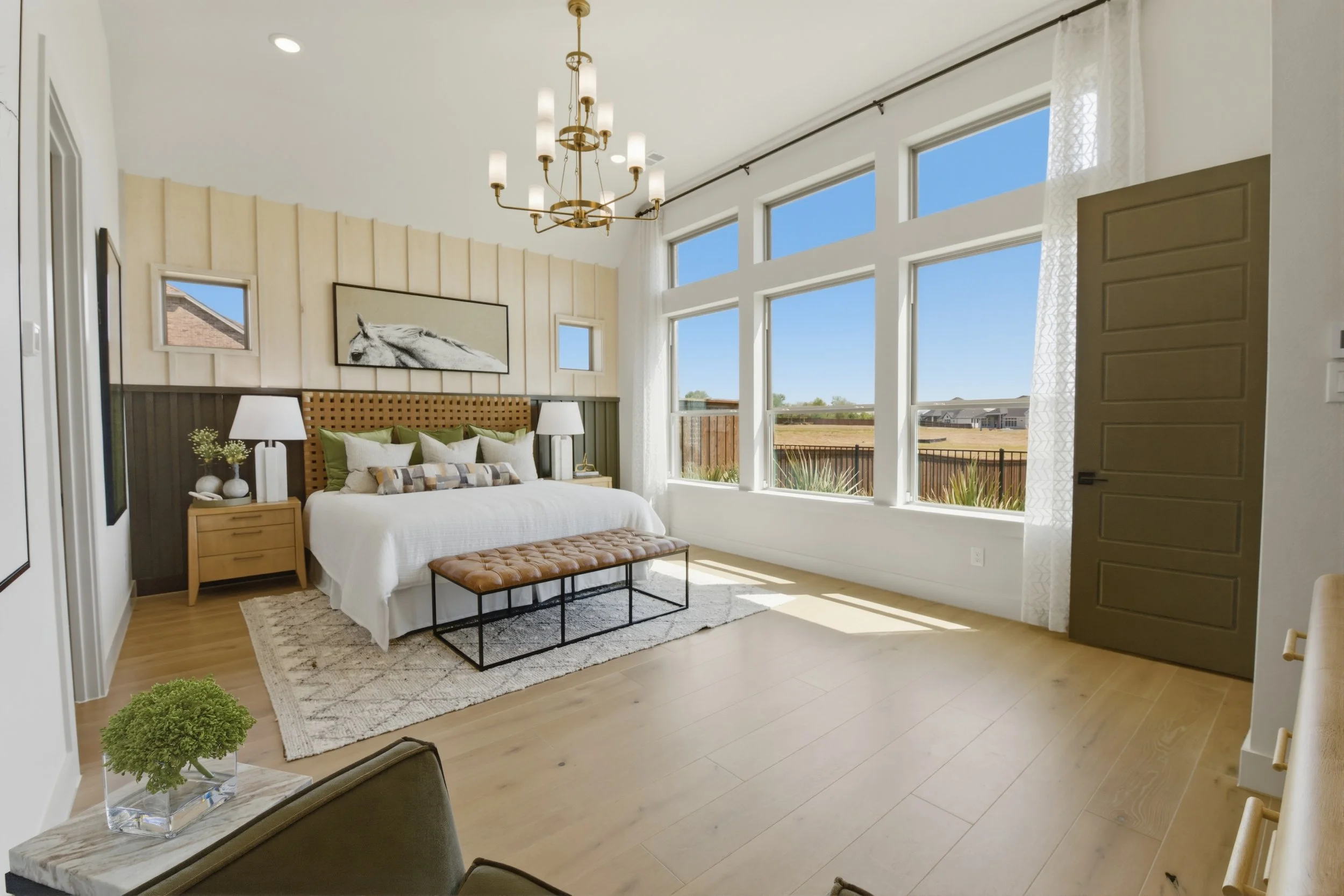 Bright bedroom with large windows, wooden floors, a bed with green and white pillows, a brown tufted bench, two nightstands with lamps, wall art of a horse, and minimal decor.
