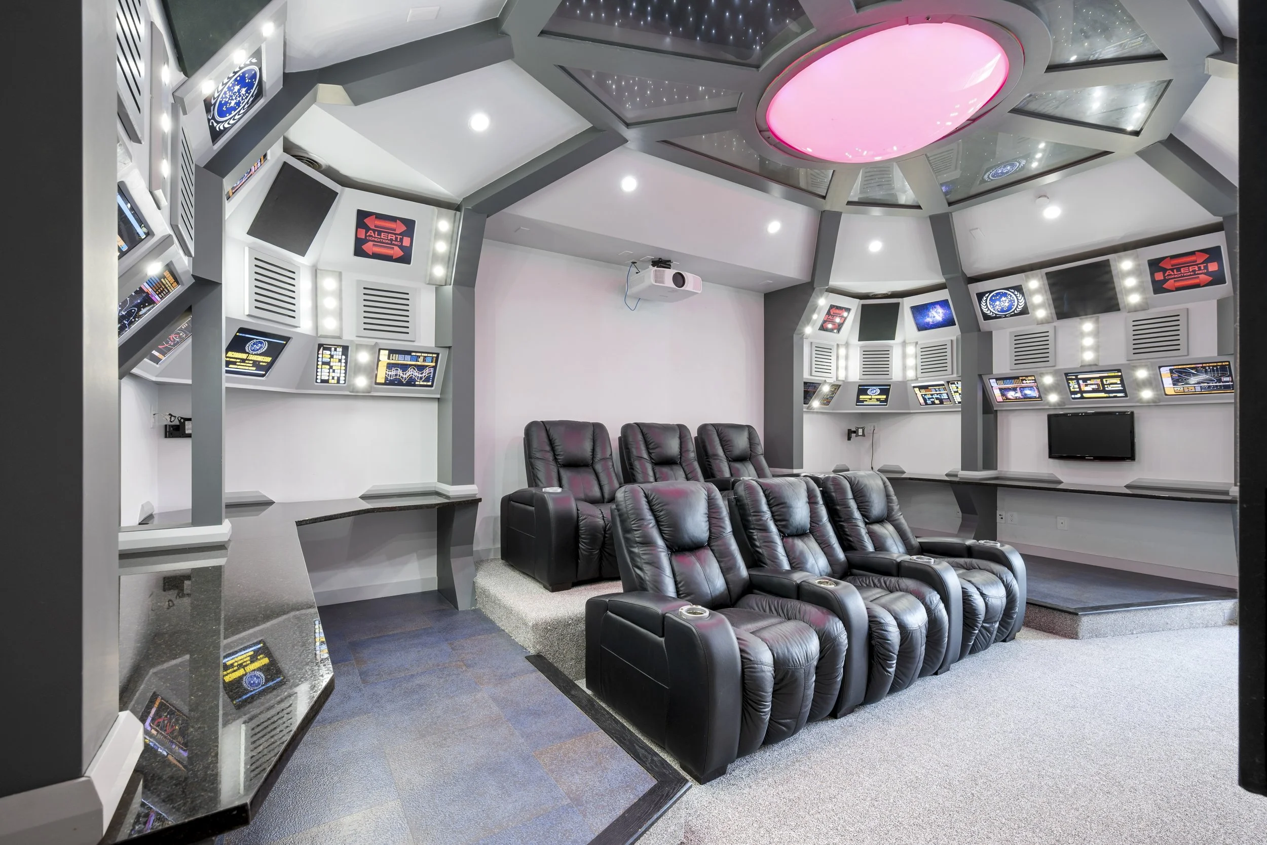 A futuristic home theater or spaceship simulation room with a curved ceiling, multiple screens displaying space graphics, and black leather recliner seats arranged in rows.
