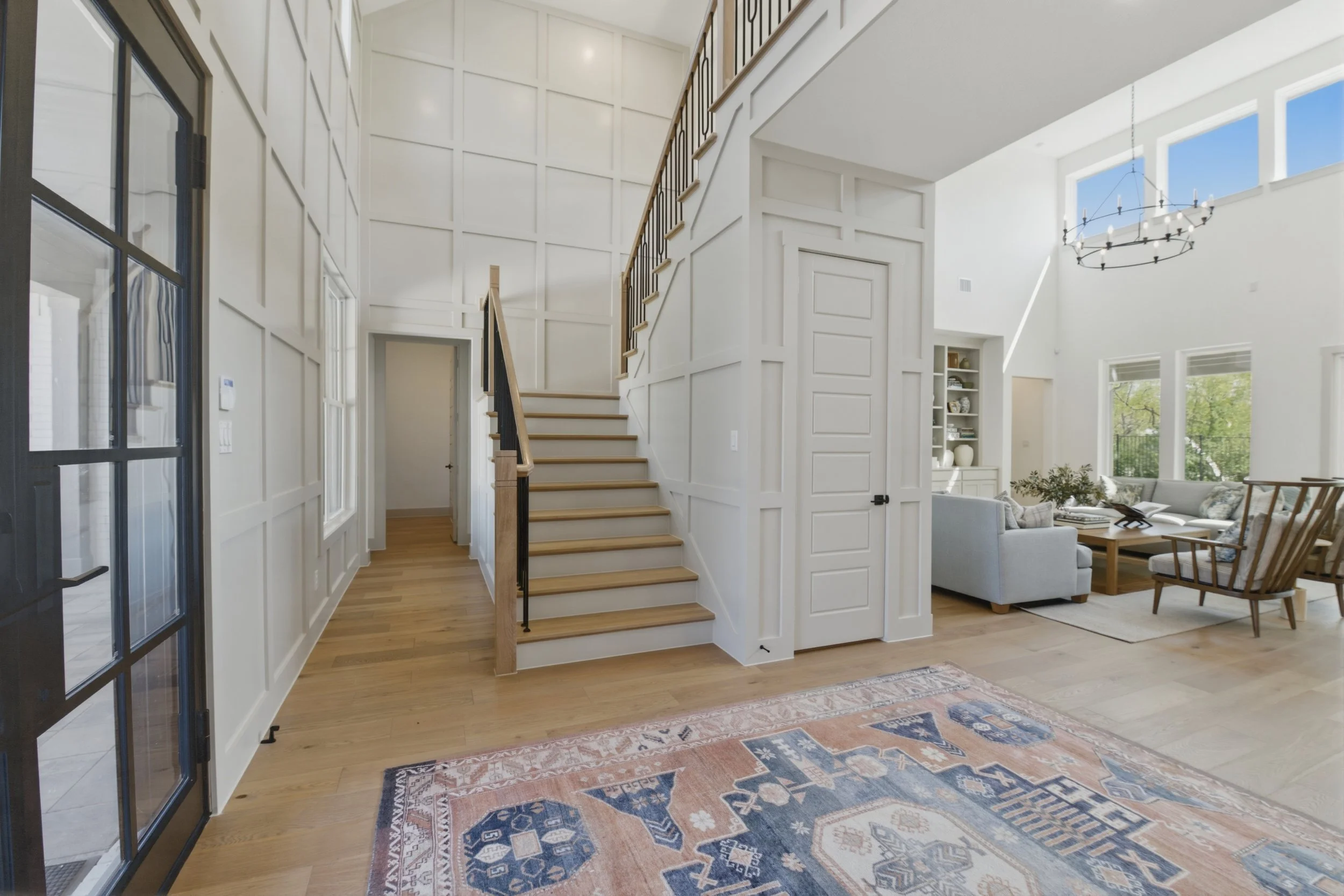 Interior view of a two-story house with white walls, a wooden staircase, and large windows. The space includes a cozy sitting area with a white sofa, wooden armchairs, and a patterned rug. A black chandelier hangs from the ceiling.