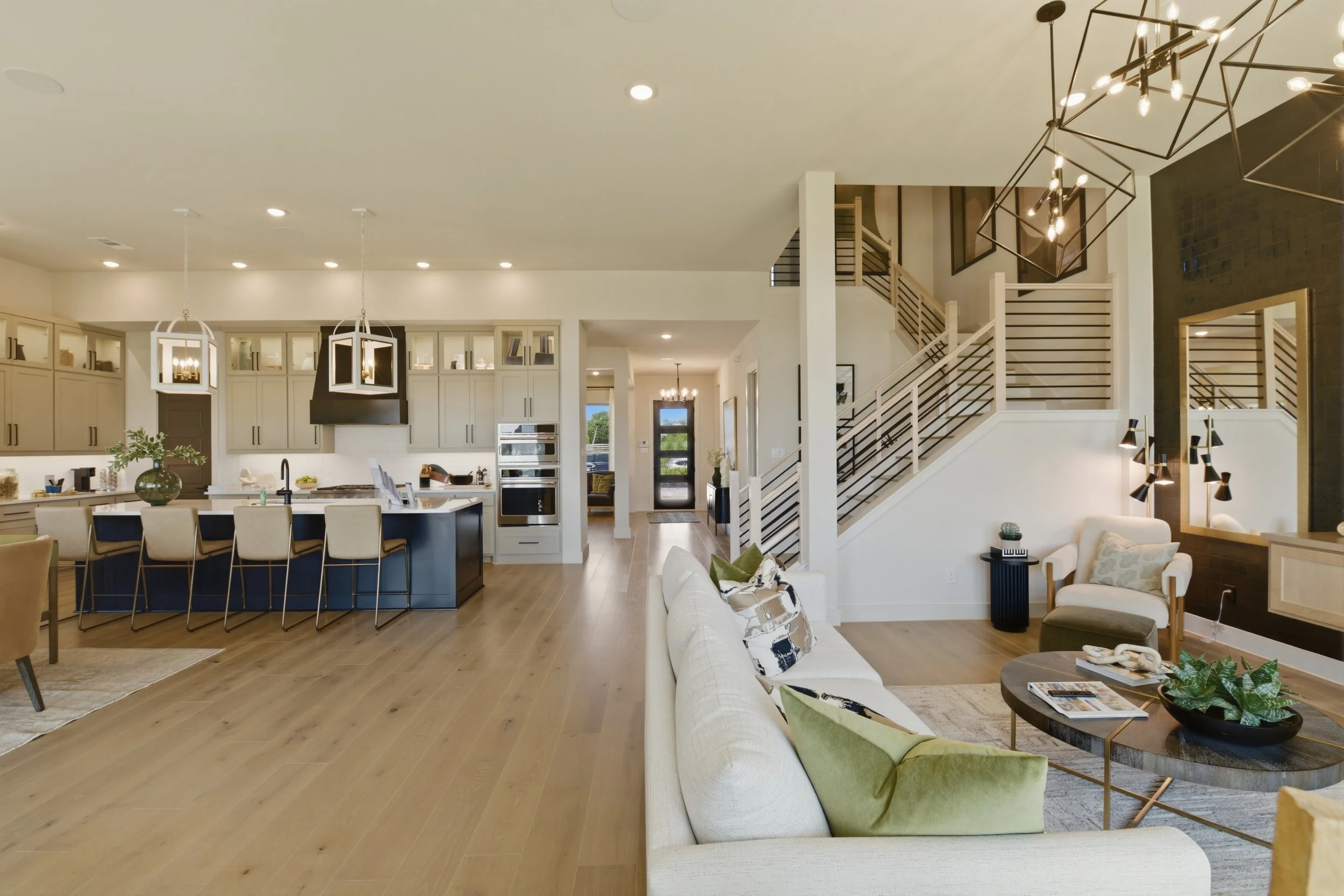 Open-concept kitchen and living room with modern decor, hardwood floors, a kitchen island with barstools, white cabinetry, stylish lighting fixtures, and a staircase leading upstairs.