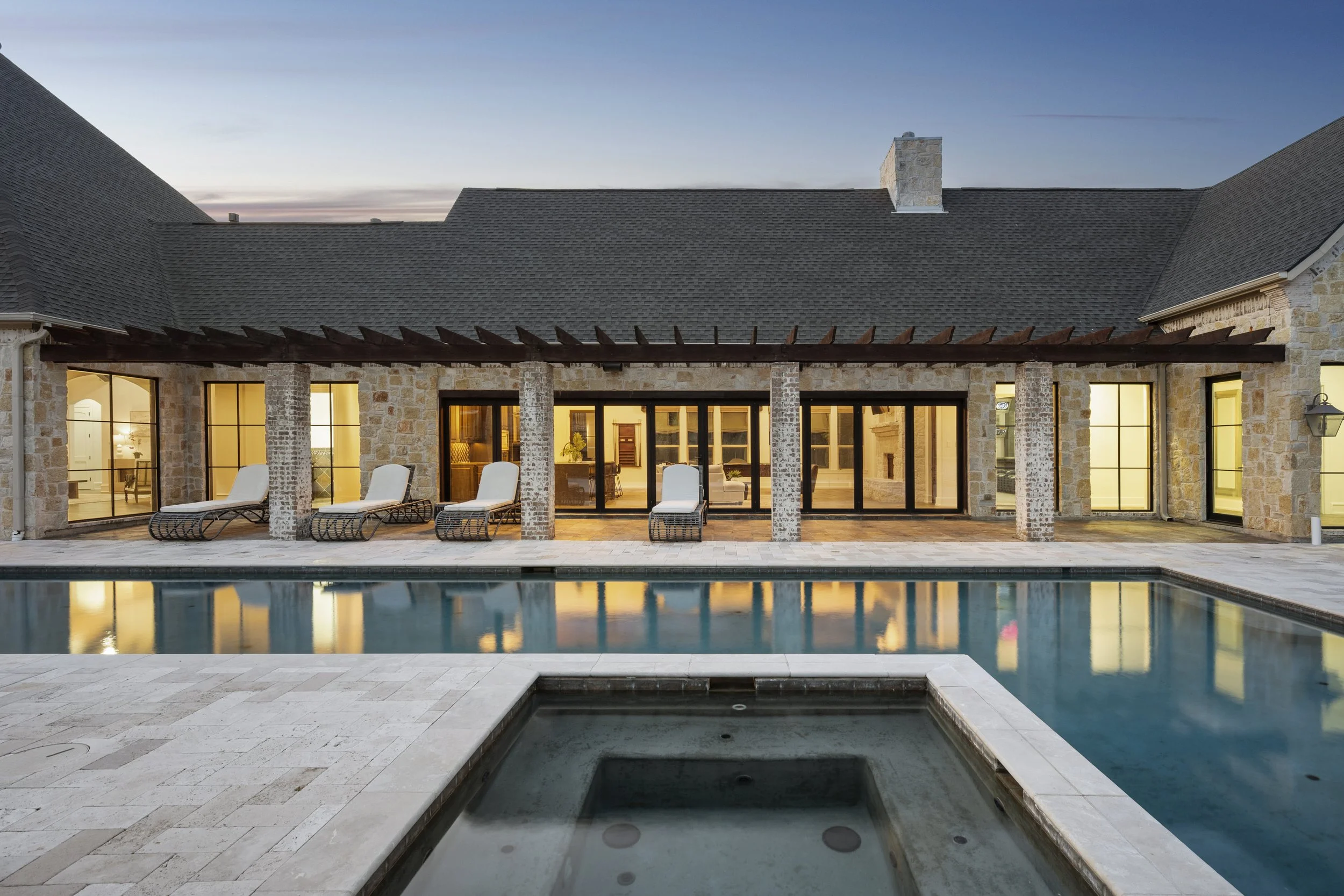 View of a backyard with a swimming pool, lounge chairs, large glass doors, and a stone house with a pitched roof at dusk.