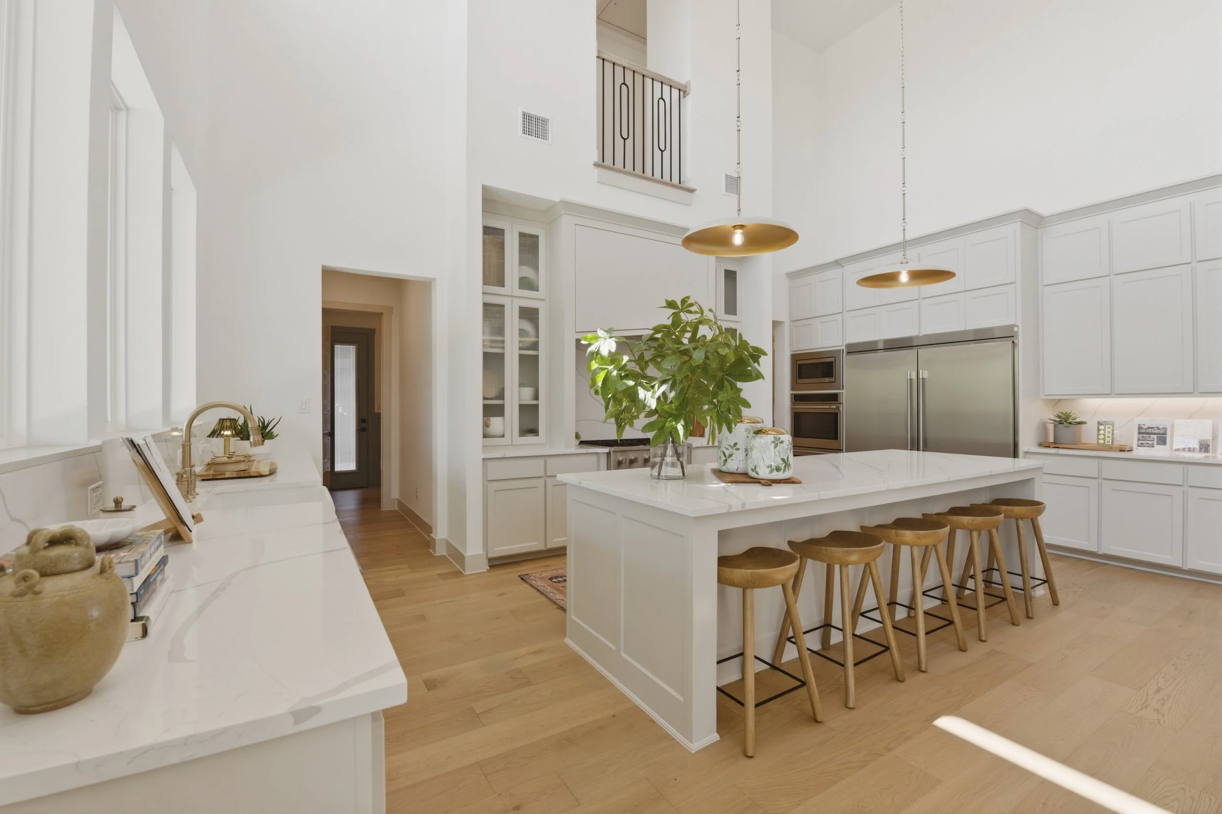 Bright, modern kitchen with white cabinets, a large island with seating, pendant lights, wooden stools, and a stainless steel refrigerator. There are green plants and decorative items on the countertops.