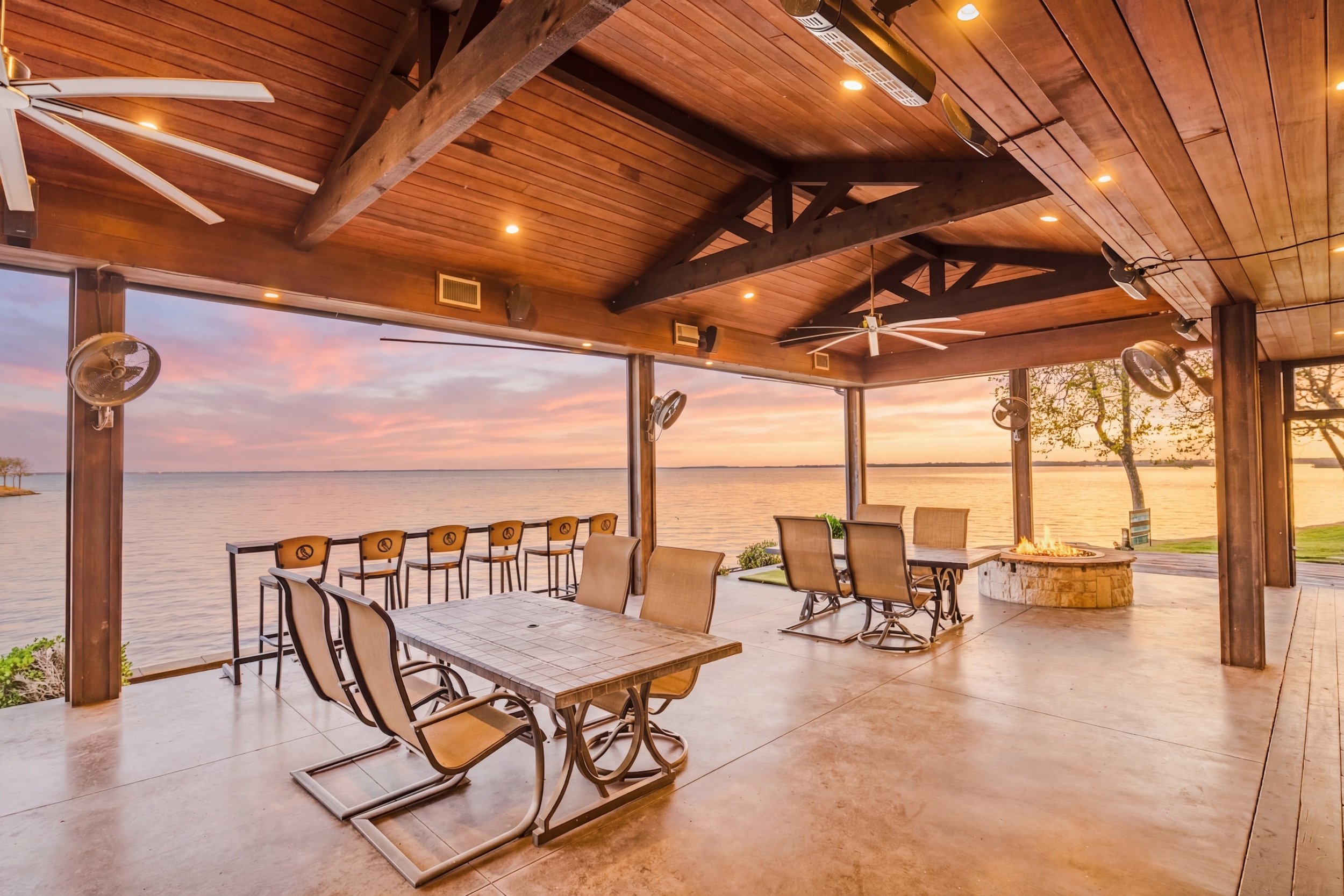 A lakeside outdoor patio with wooden ceiling beams, ceiling fans, and a firepit, overlooking a calm lake at sunset.