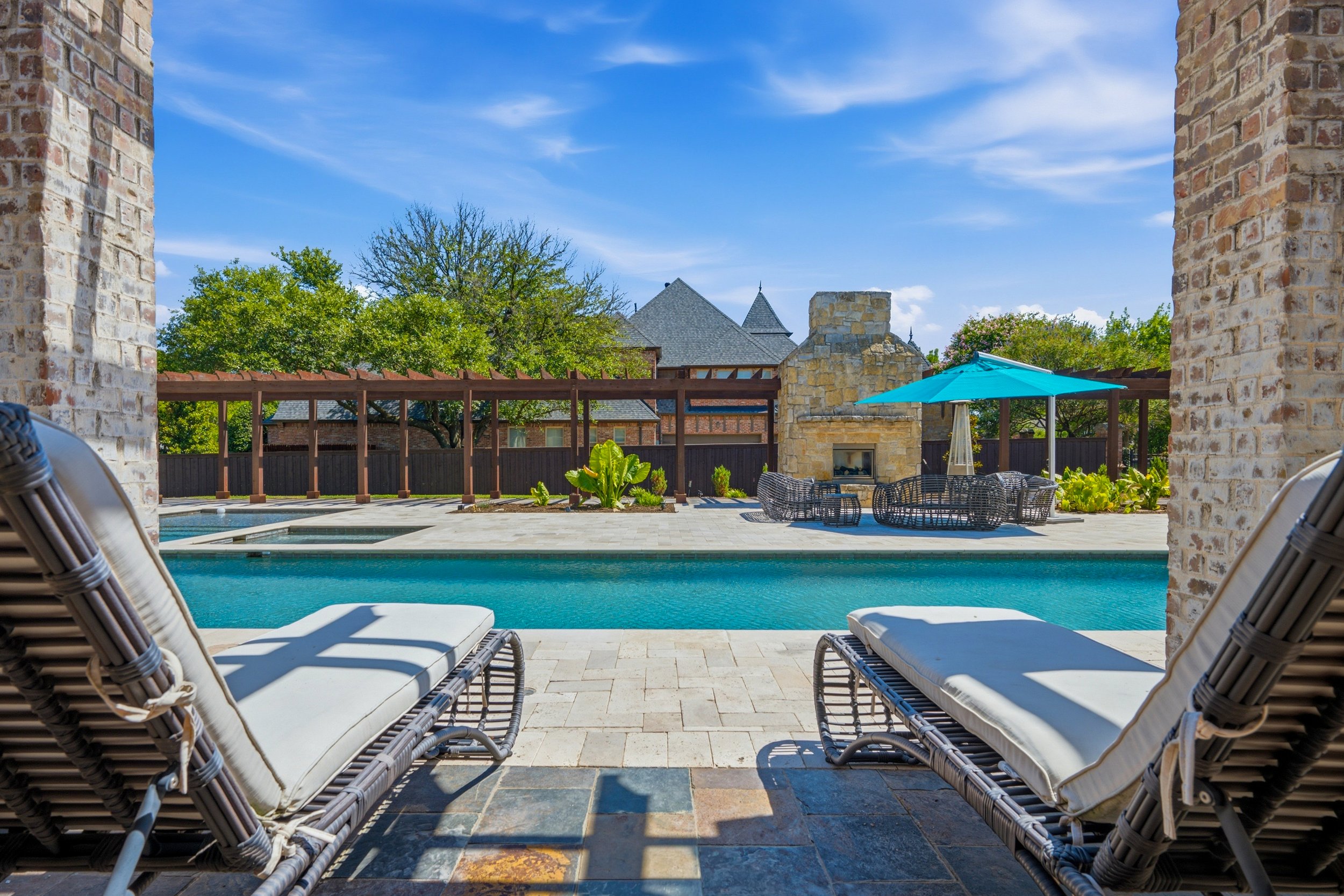 View of a backyard pool area framed by two brick columns, with lounge chairs in the foreground, a covered patio, and a stone fireplace with a patio umbrella in the background under a bright blue sky.