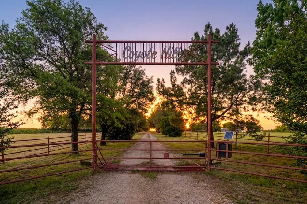 Entrance gate to a private property, with a sign reading 'Coyote Hill', leading to a dirt pathway flanked by wooden fences and trees at sunset.