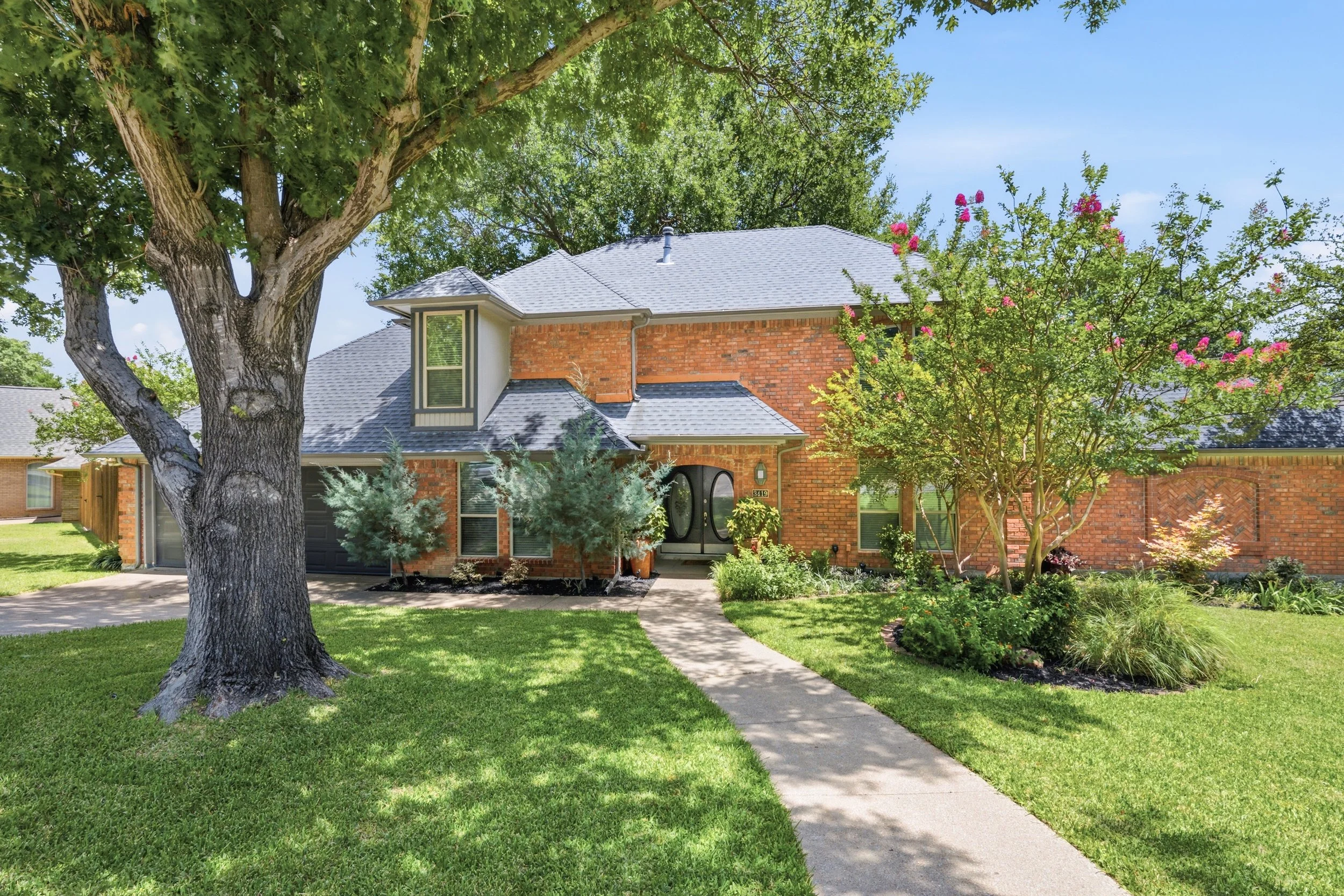 Front yard of a brick house with a tree on the left and flowering shrub on the right, a sidewalk leading to the front door, and a well-maintained lawn under a blue sky.
