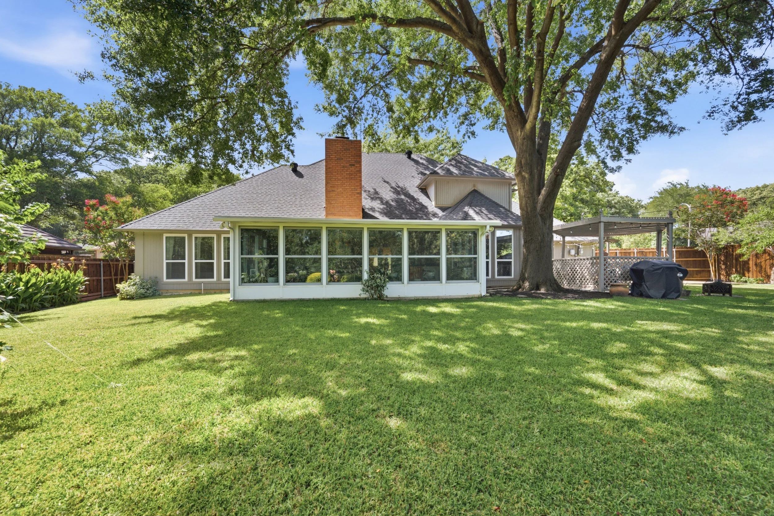 Lawn backyard with large tree, house with screened porch, and fenced yard on sunny day.