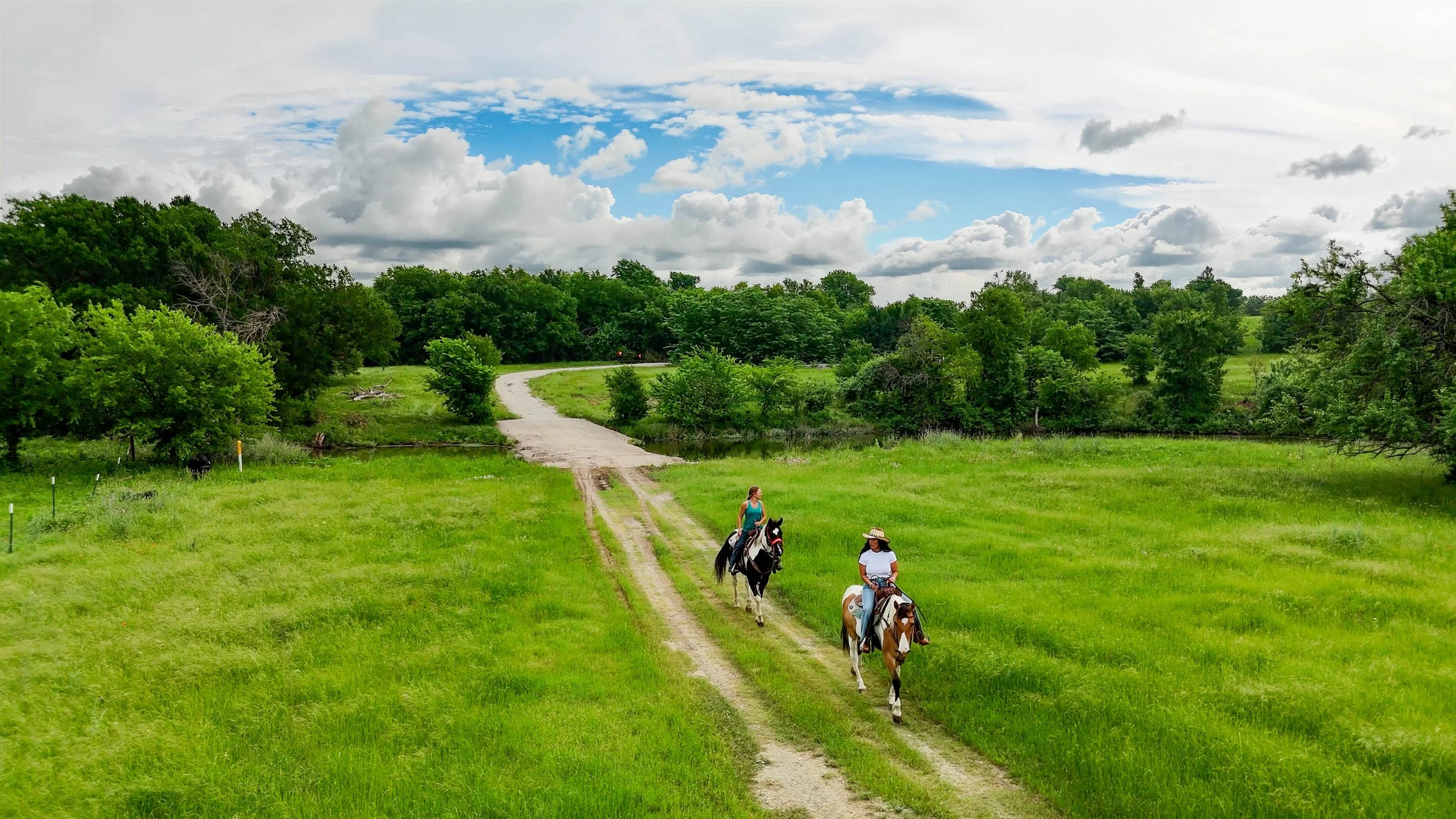 Two people riding horses along a dirt path in a lush green field, with dense trees and a cloudy sky in the background.