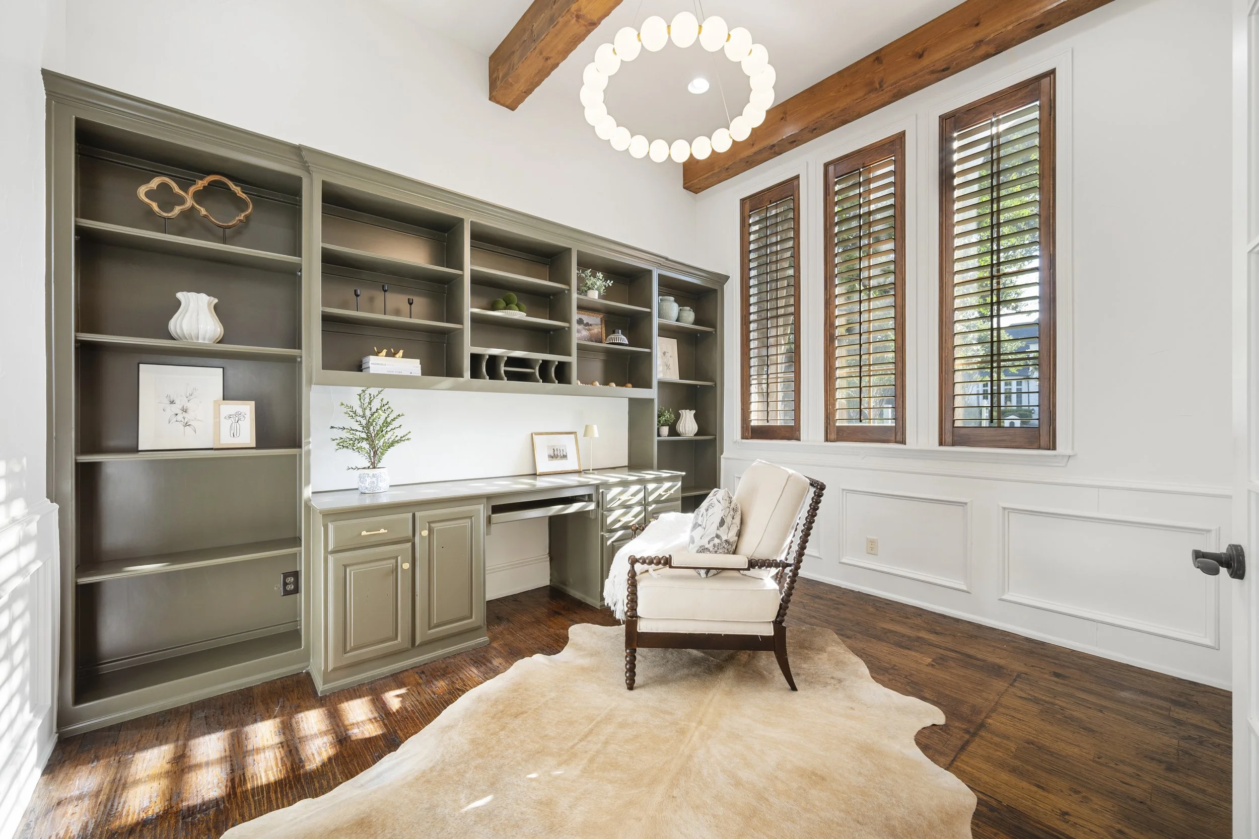 A cozy living room corner with a green built-in bookcase, a white cushioned armchair, a wooden ceiling beam, large windows with wooden shutters, and a circular chandelier hanging from the ceiling.