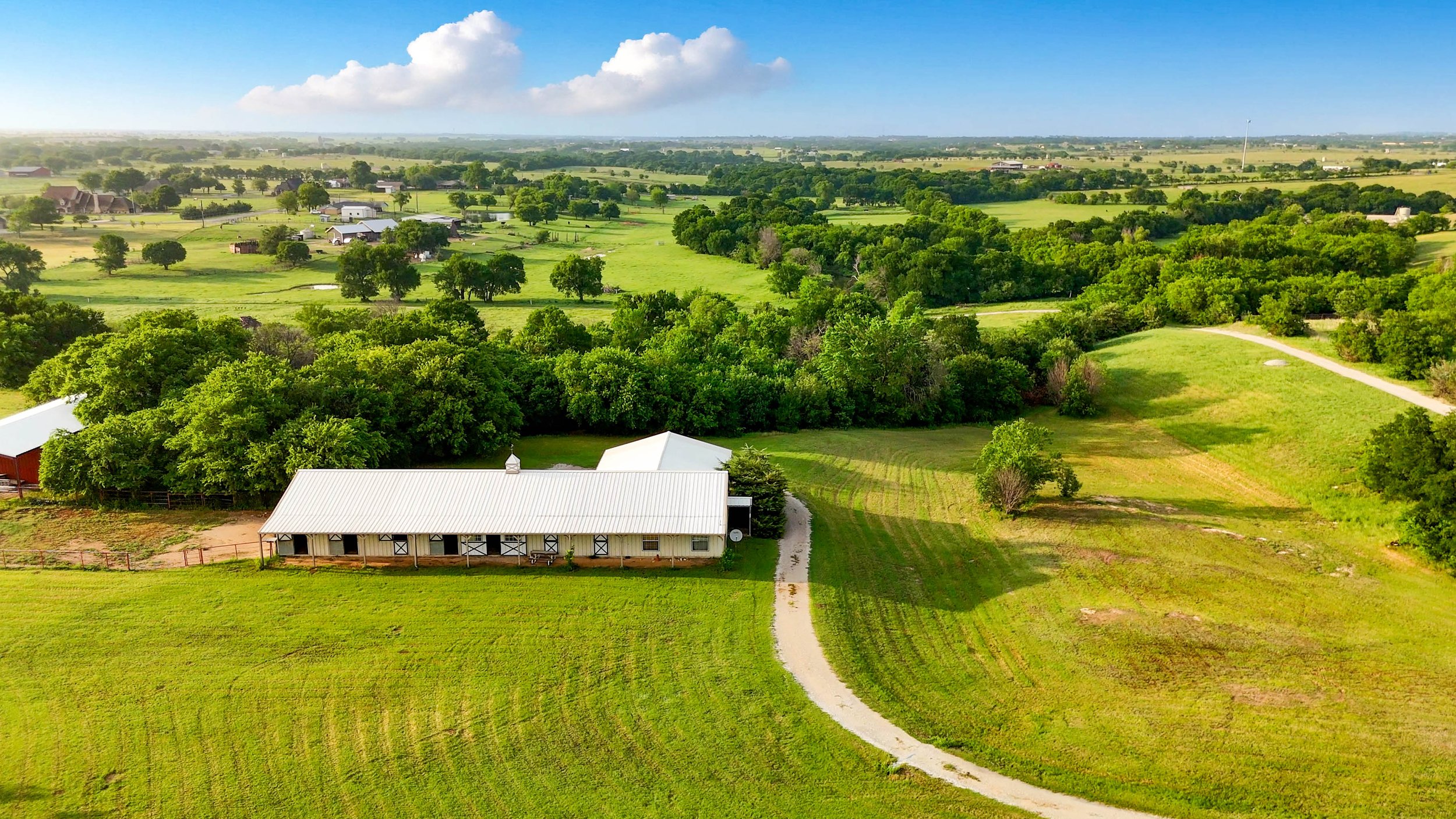 Aerial view of a farm with a white barn, green fields, trees, and a winding gravel driveway.