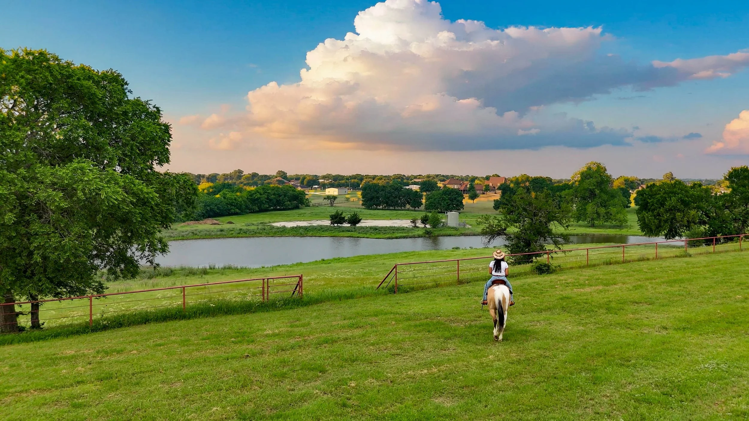 Person riding a horse along a grassy area near a body of water, with trees, houses, and a partly cloudy sky in the background.