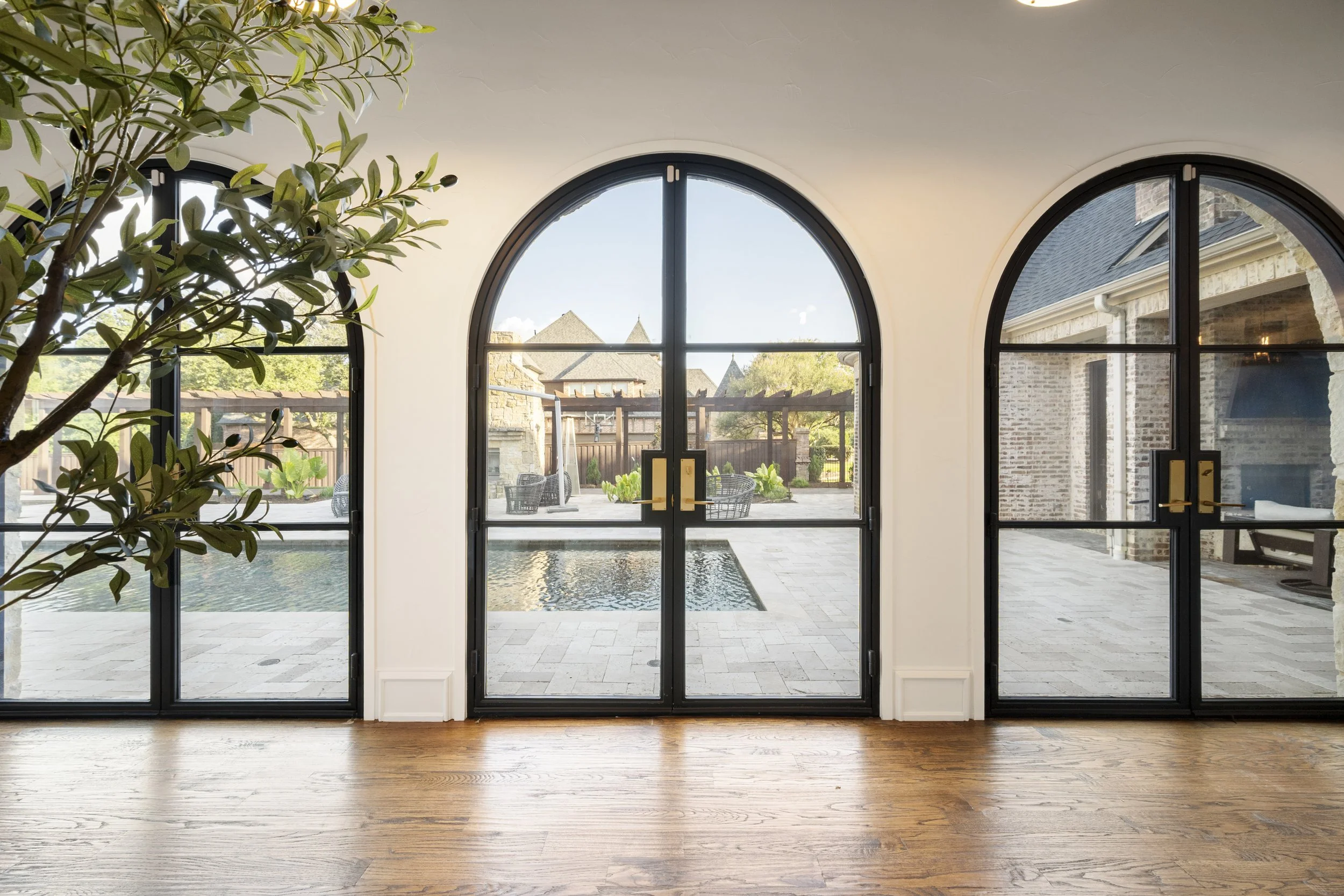 Interior view of a room with three large arched glass doors leading outside to a patio and backyard with trees, fencing, and neighboring houses visible. The room has wooden flooring and a large potted plant on the left.