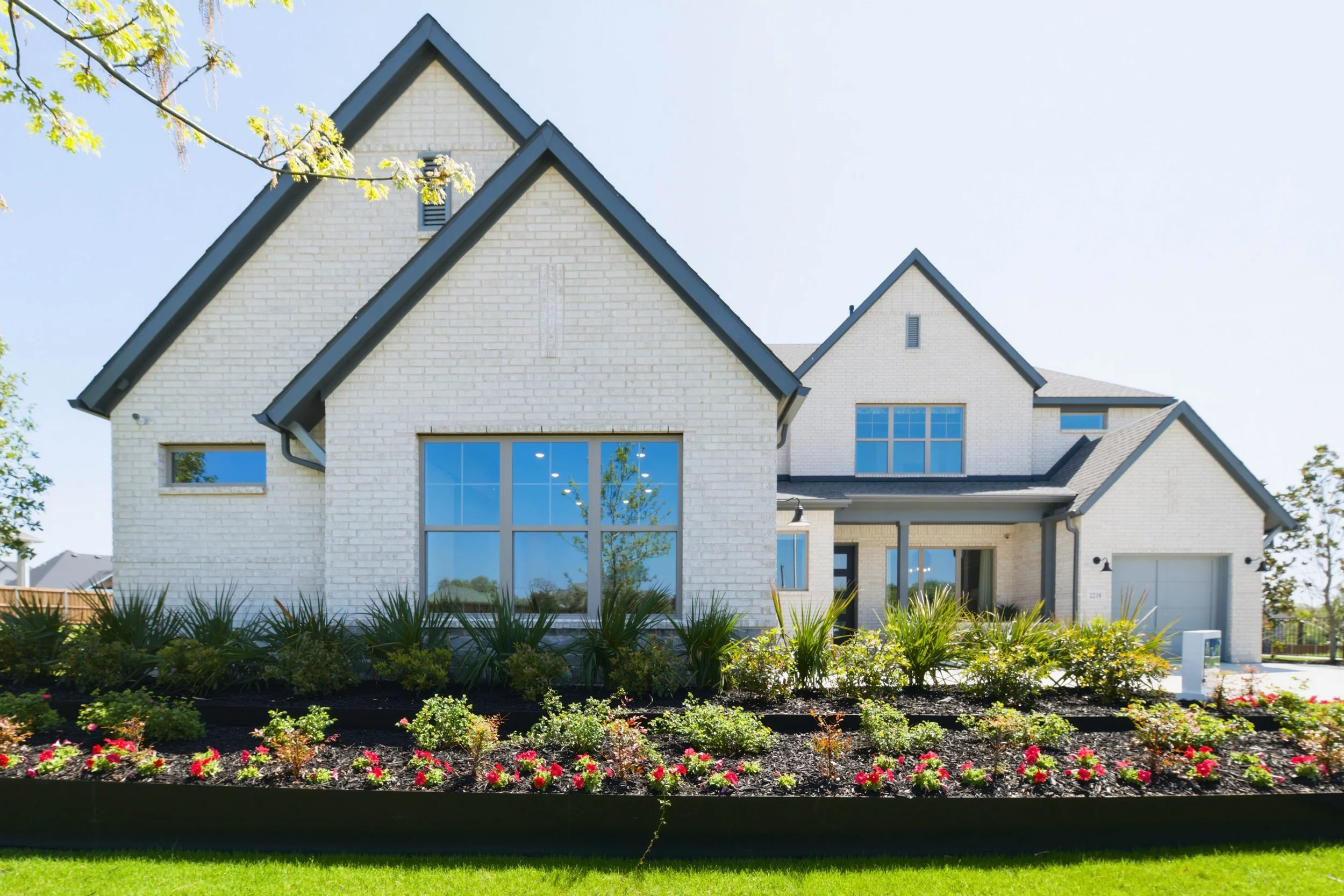 A modern house with white brick exterior and large windows, surrounded by a well-maintained garden with colorful flowers and green grass, under a clear blue sky.