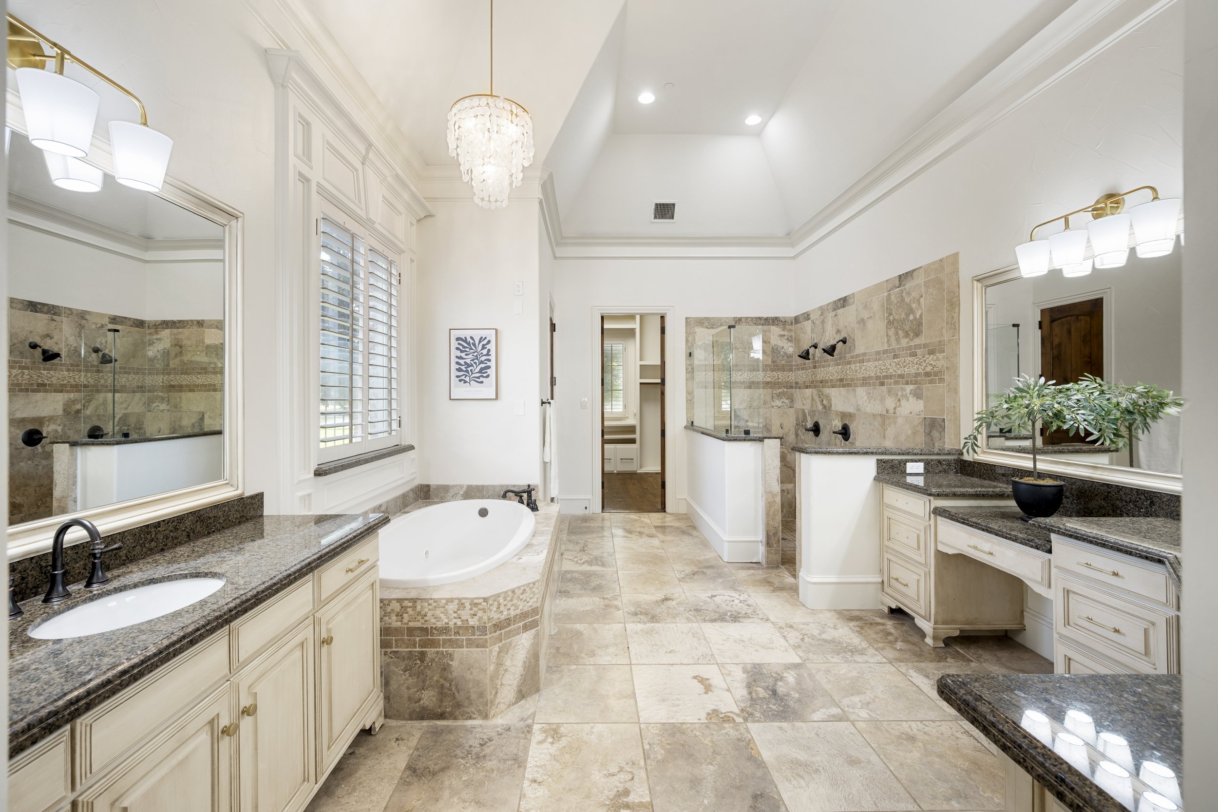 Bright bathroom with white walls, beige tile floors, and a large bathtub. Features include double vanities with granite countertops, large mirrors, and wall-mounted light fixtures. There's a window with plantation shutters, a chandelier, and a walk-i