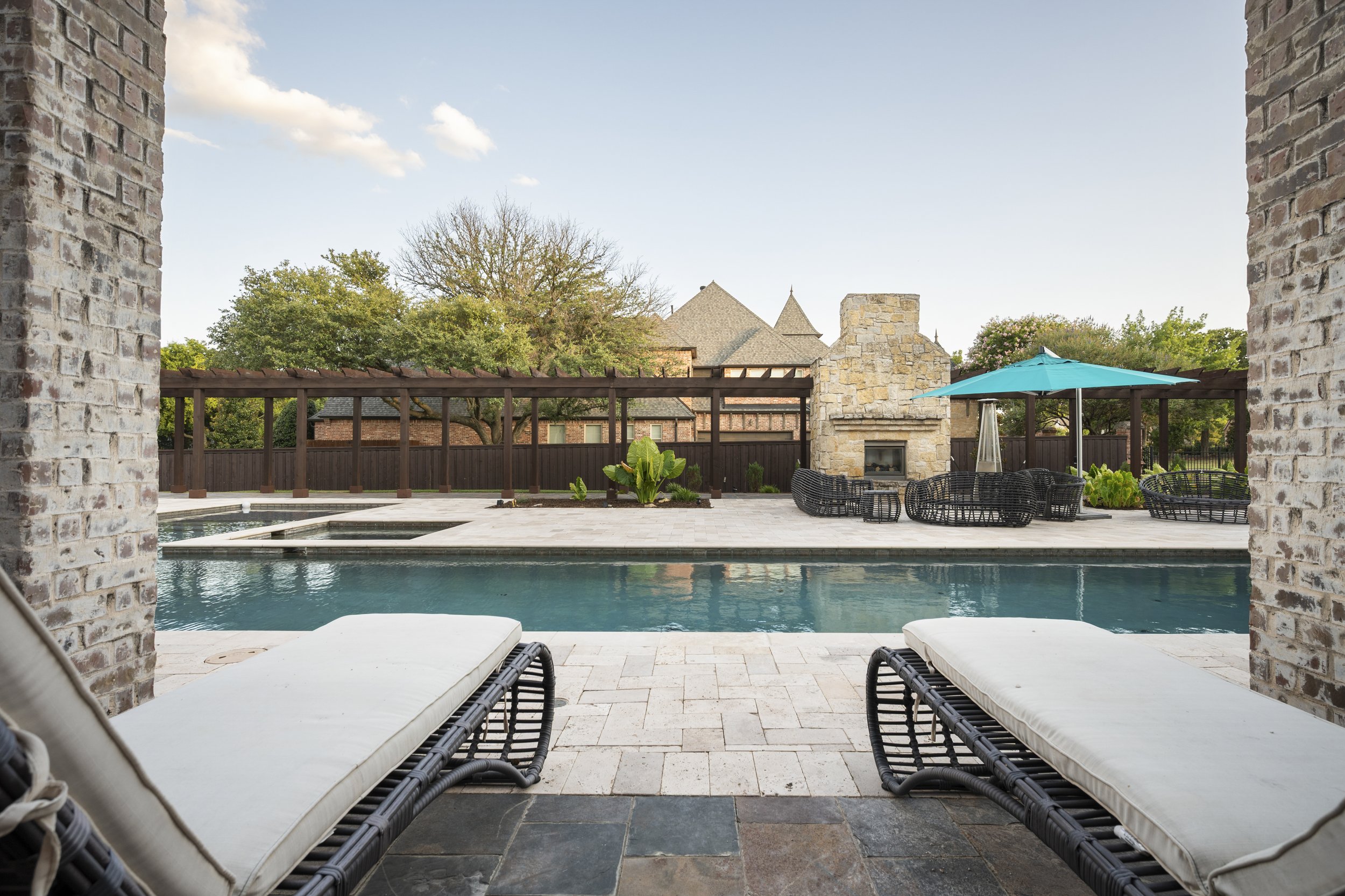 View of a backyard with a swimming pool, surrounded by a stone patio and outdoor furniture including lounge chairs, a blue patio umbrella, and a fireplace with stone design. The background features a wooden trellis, trees, and a house with a pitched 