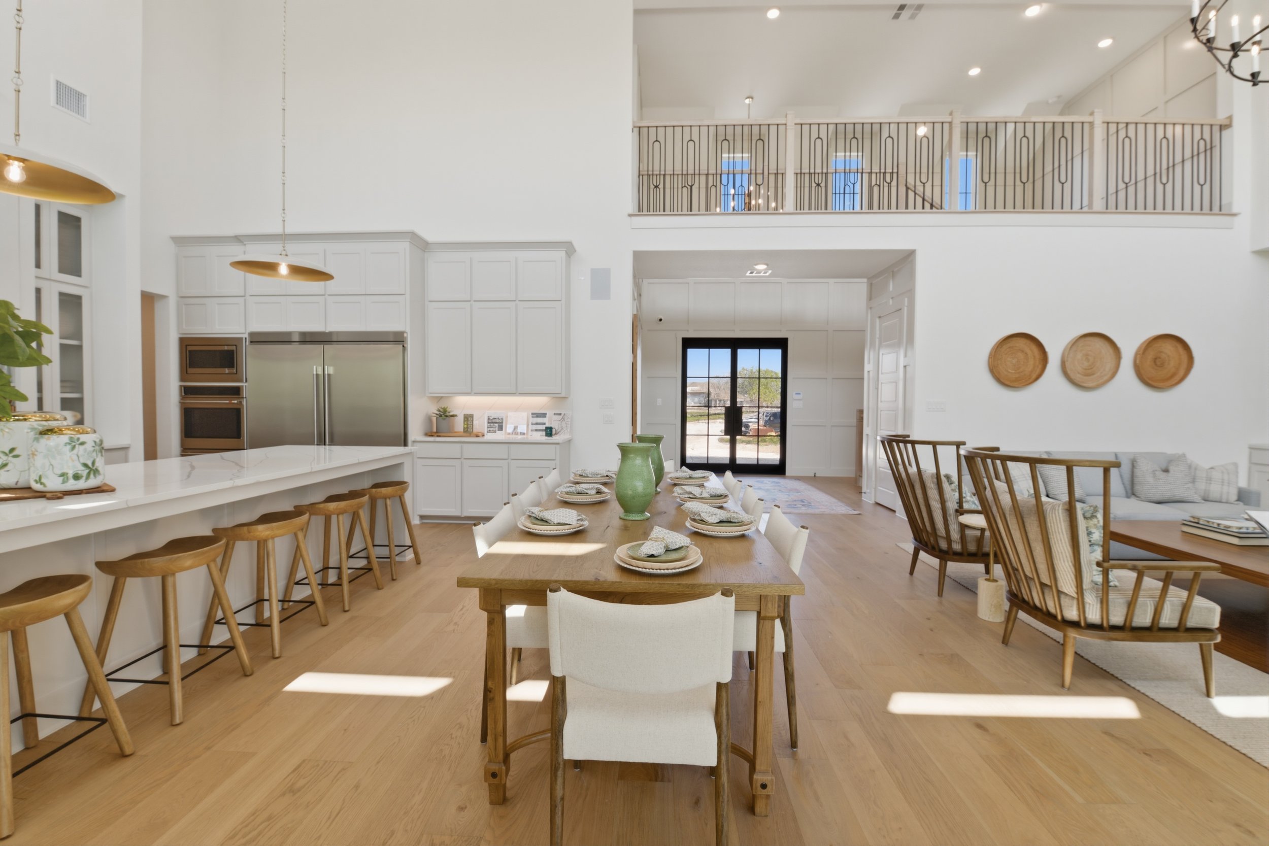Open-concept kitchen and dining area with white cabinets, stainless steel appliances, dining table with tableware, and a sitting area with chairs and a sofa, high ceiling with chandelier, and a balcony railing.