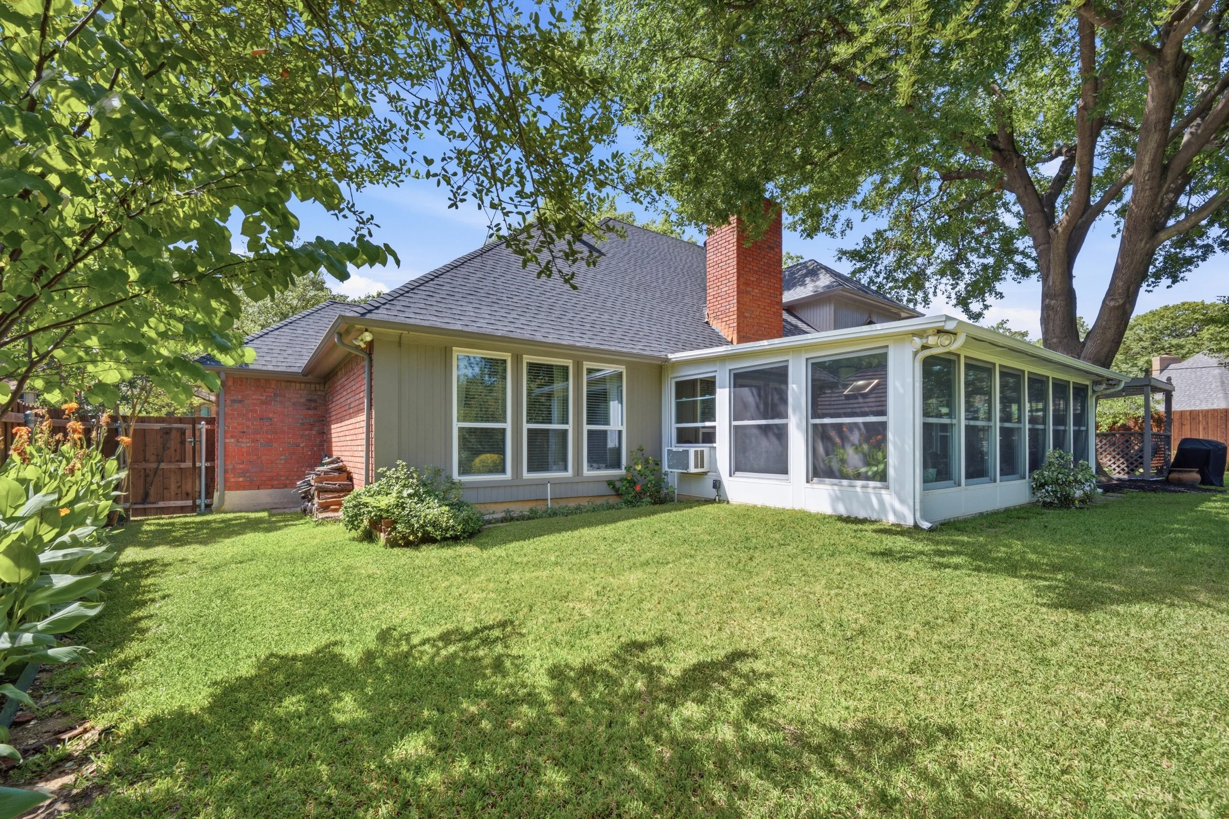 Backyard view of a house with large windows, a brick chimney, and a screened porch, surrounded by green grass, trees, and shrubs under a blue sky.