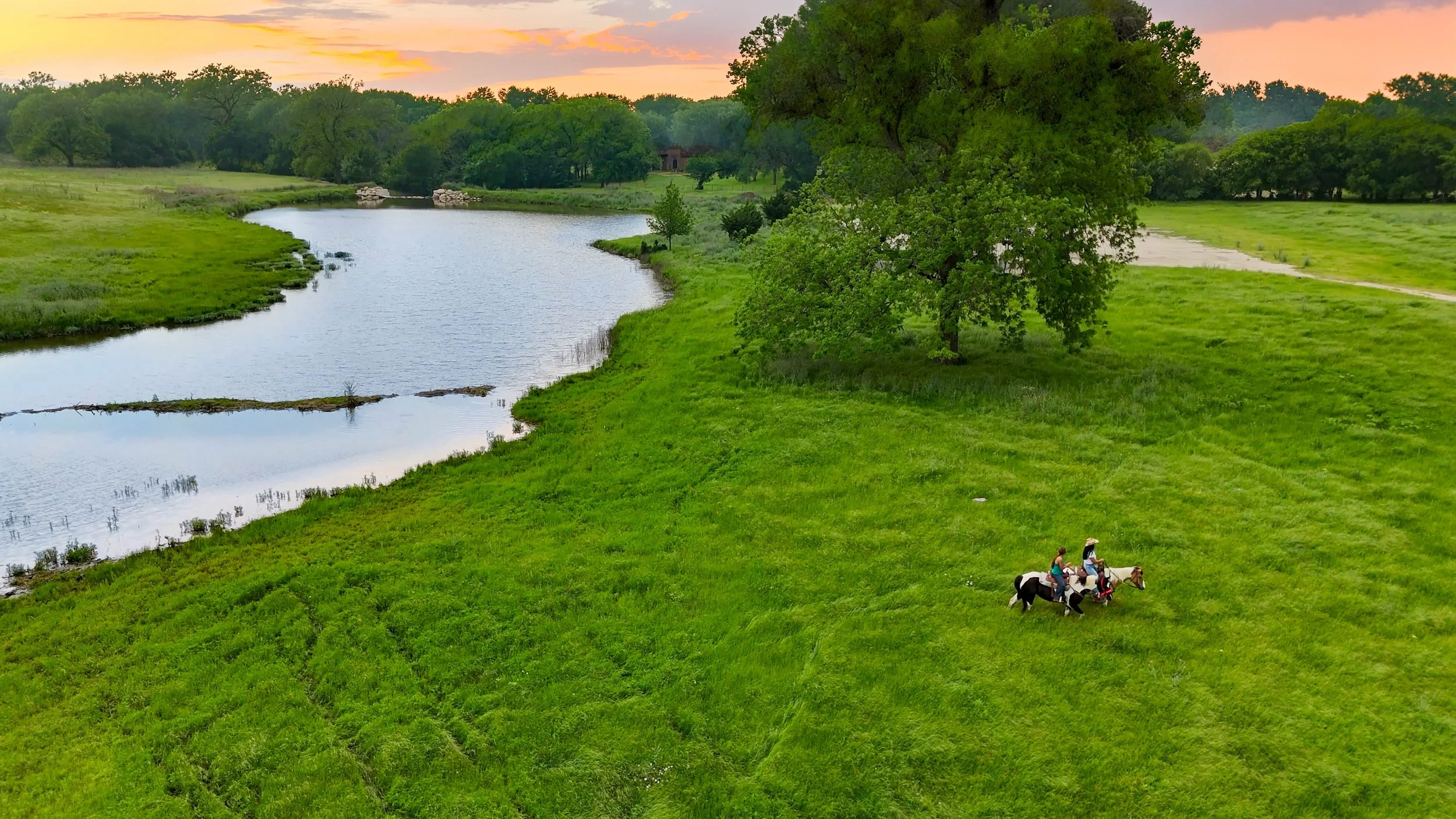 A scenic landscape with a river running through lush green grass and trees at sunset; two people horseback riding in a field.