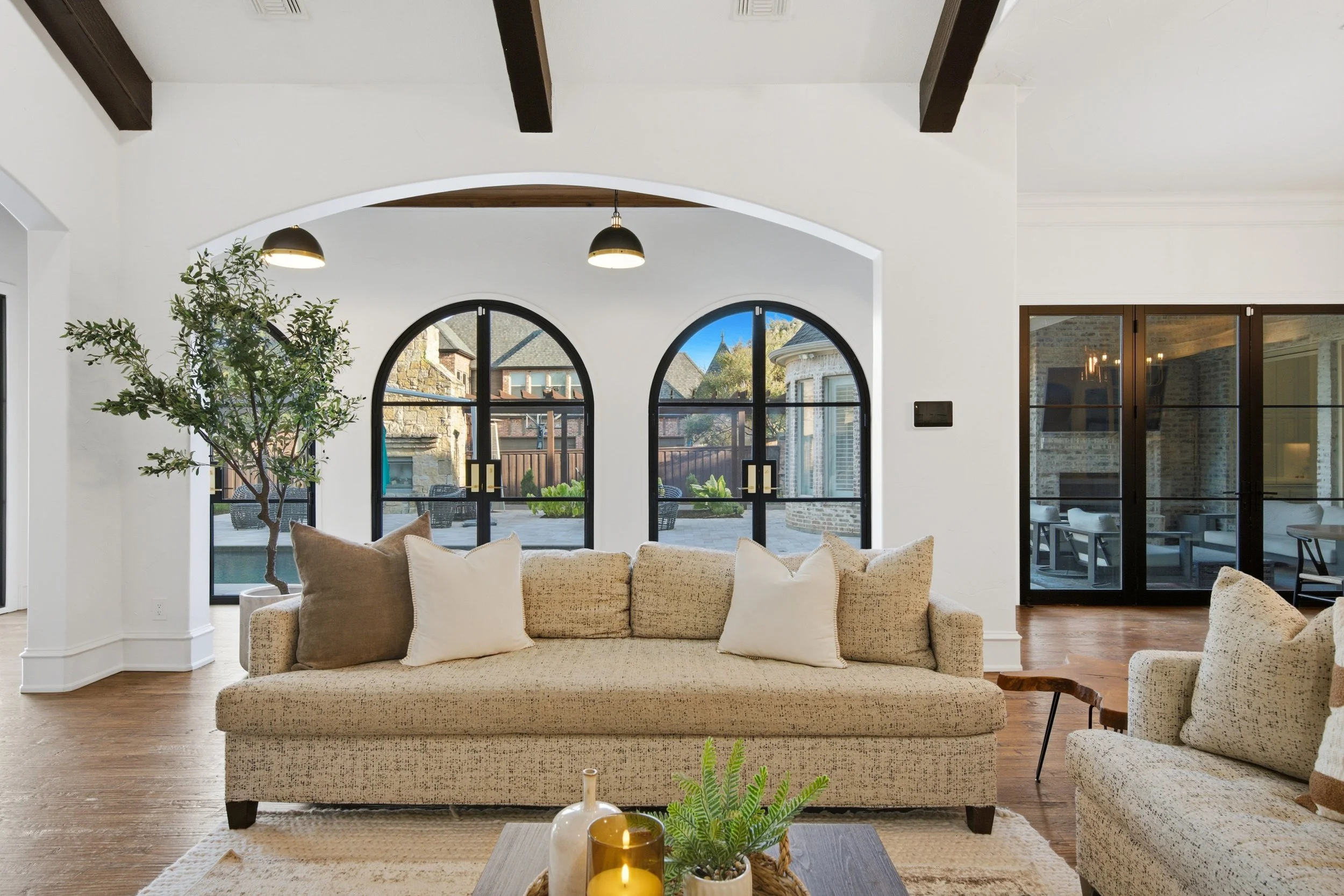 A spacious living room with a beige sofa, decorative pillows, and a coffee table with candles and plants. Large arched windows overlook a backyard with patio furniture and neighboring houses. White walls, wooden flooring, and ceiling beams add to the