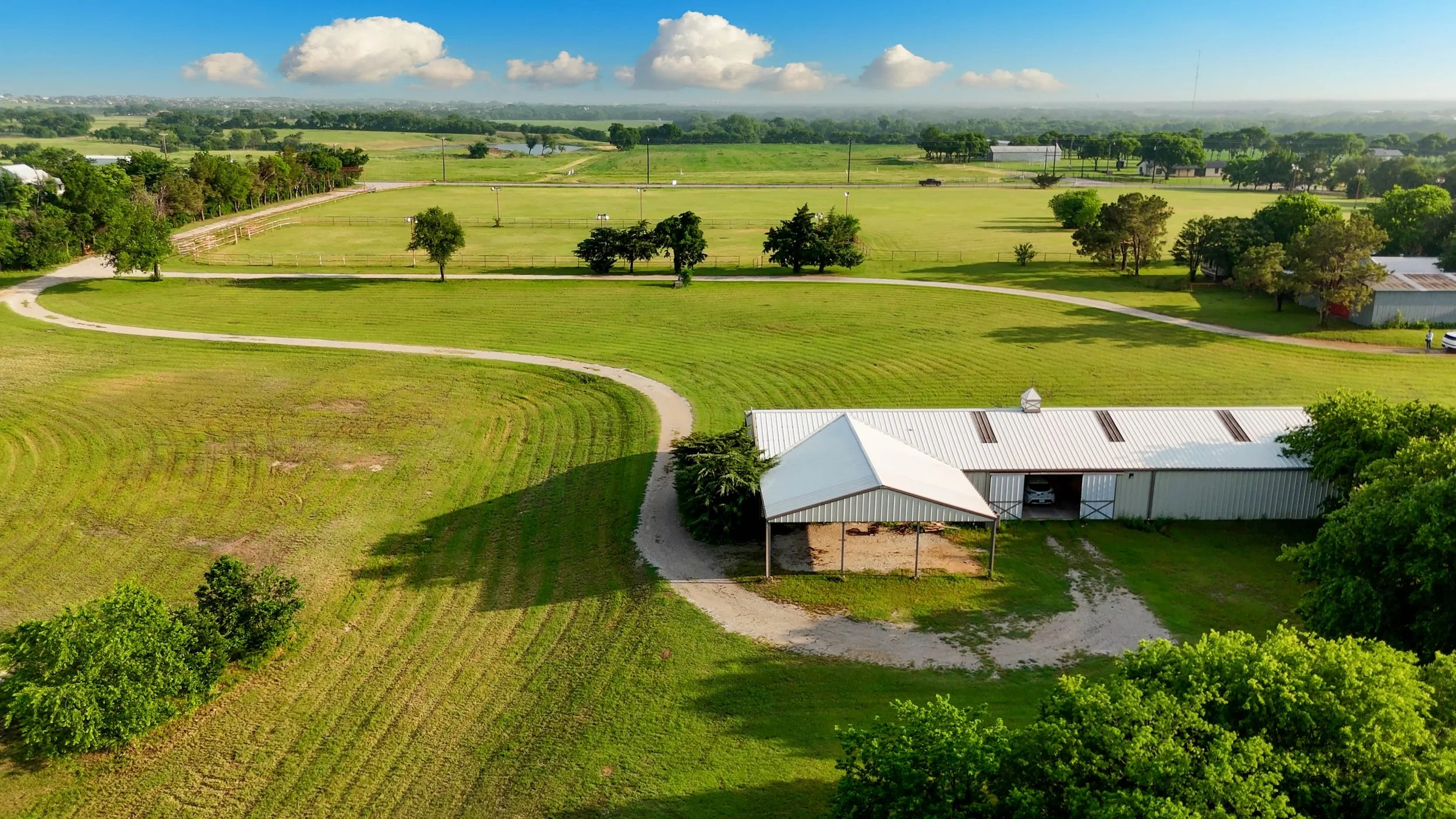 Aerial view of a large green farm with open fields, trees, a few buildings, and a winding dirt path on a sunny day with a blue sky and scattered clouds.