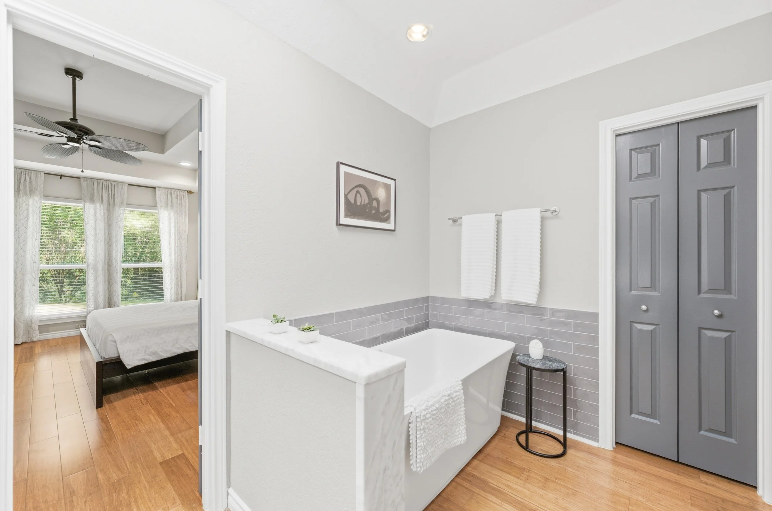 Bathroom with a bathtub, white towels on a rail, a small round black table, gray closet doors, light gray and white walls, and a framed picture. Adjacent bedroom with large windows, white curtains, and a ceiling fan.