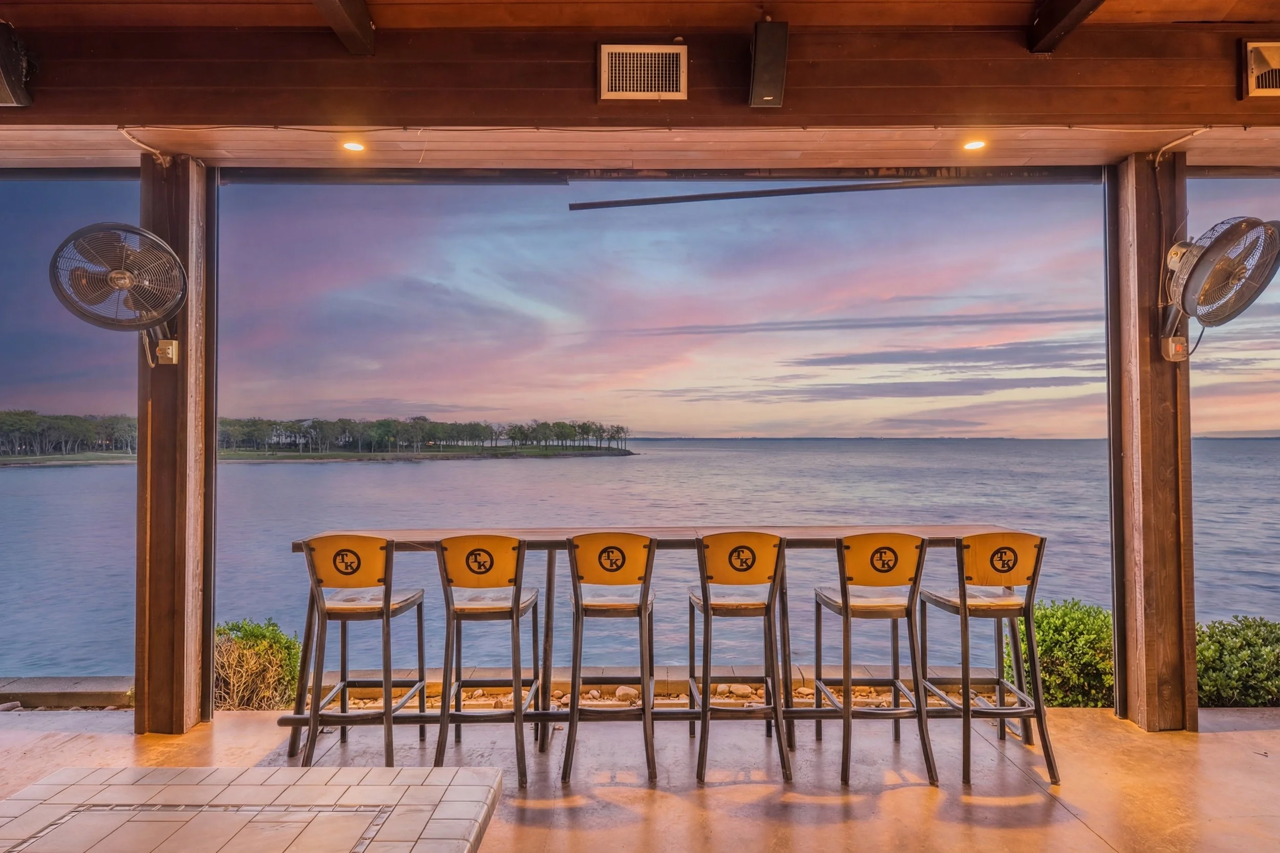 View from a covered patio overlooking a body of water with a line of green trees in the distance and a colorful sunset sky. There are six yellow chairs with black lettering on a deck, and two wall-mounted fans on either side of the open space.