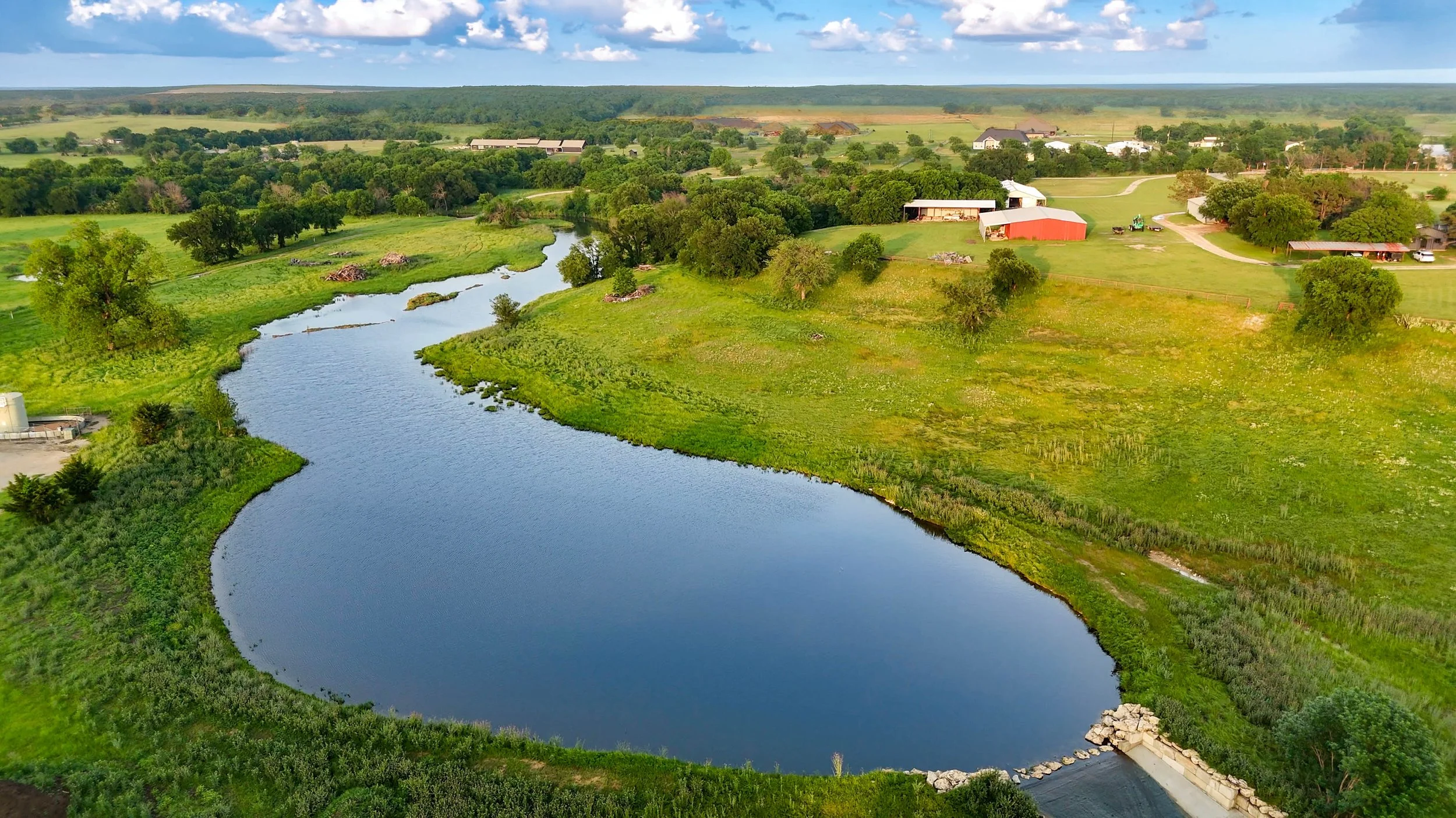 Aerial view of a green rural landscape with a winding river, trees, fields, and scattered farm buildings under a partly cloudy sky.